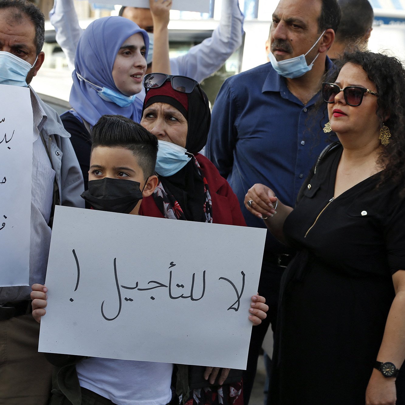 En la imagen se puede ver a un grupo de personas reunidas, algunas de las cuales sostienen carteles. La mayoría llevan mascarillas. Entre las personas, un niño sostiene un cartel que tiene un mensaje. Se observa una variedad de expresiones en sus rostros, reflejando una atmósfera de protesta o manifestación. La vestimenta de los participantes es variada, con algunas personas usando prendas tradicionales. El ambiente sugiere una situación de movilización social.