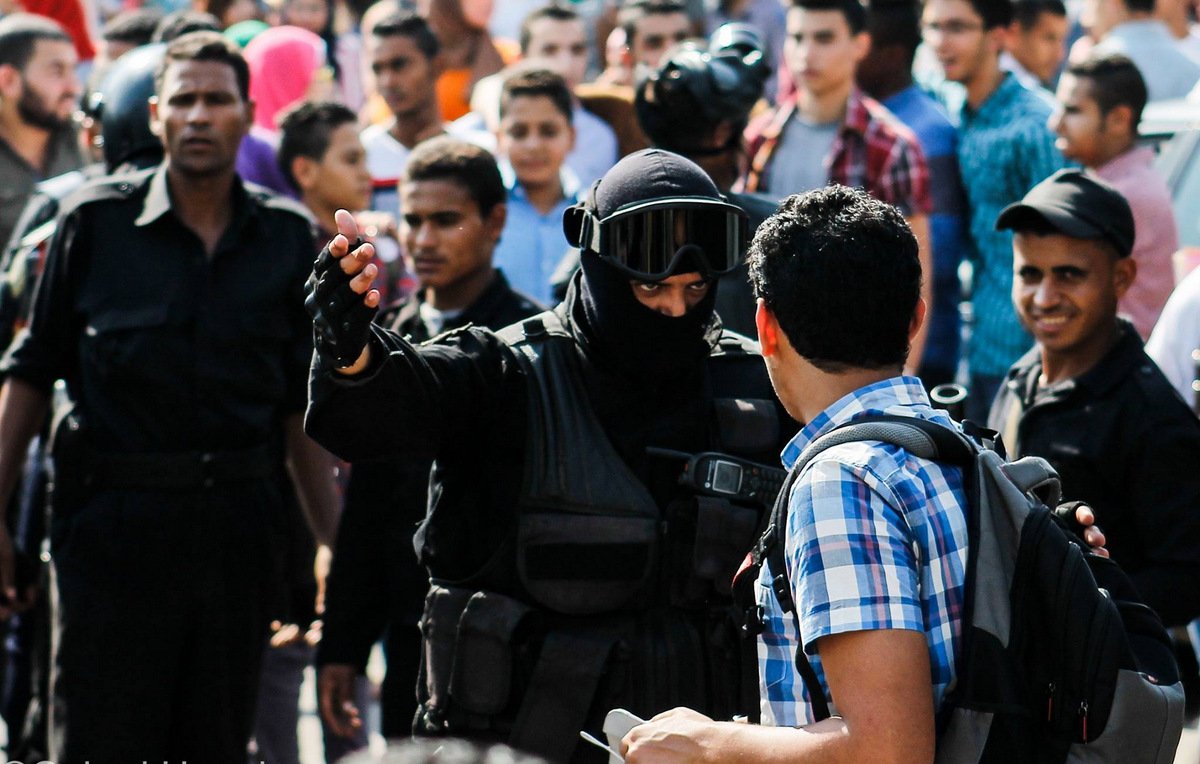 L'image montre une scène de confrontation dans un contexte urbain. À l'avant, un homme vêtu d'un uniforme noir, portant un casque et une visière, semble interagir avec un autre homme qui porte une chemise à carreaux. Ce dernier semble questionné ou arrêté, tandis qu'un groupe de personnes les entoure, observant la situation. On peut percevoir une atmosphère tendue, avec des expressions variées sur les visages des spectateurs. La scène suggère un moment de tension ou de dispute, probablement lié à une manifestation ou à une intervention policière.