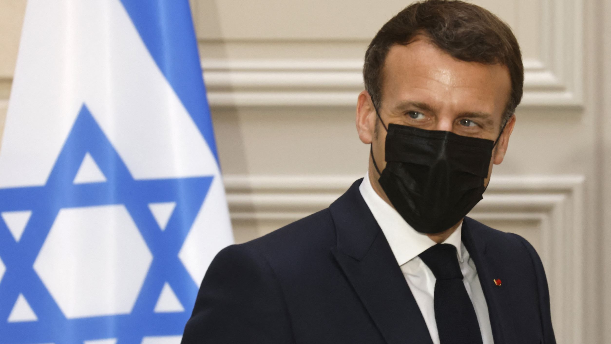 The image shows a man in a dark suit wearing a black face mask. He appears to be looking slightly to the side, with a serious expression. In the background, there is a flag featuring a blue Star of David on a white field, which likely signifies that the setting is related to Israel. The overall atmosphere suggests a formal or diplomatic context.