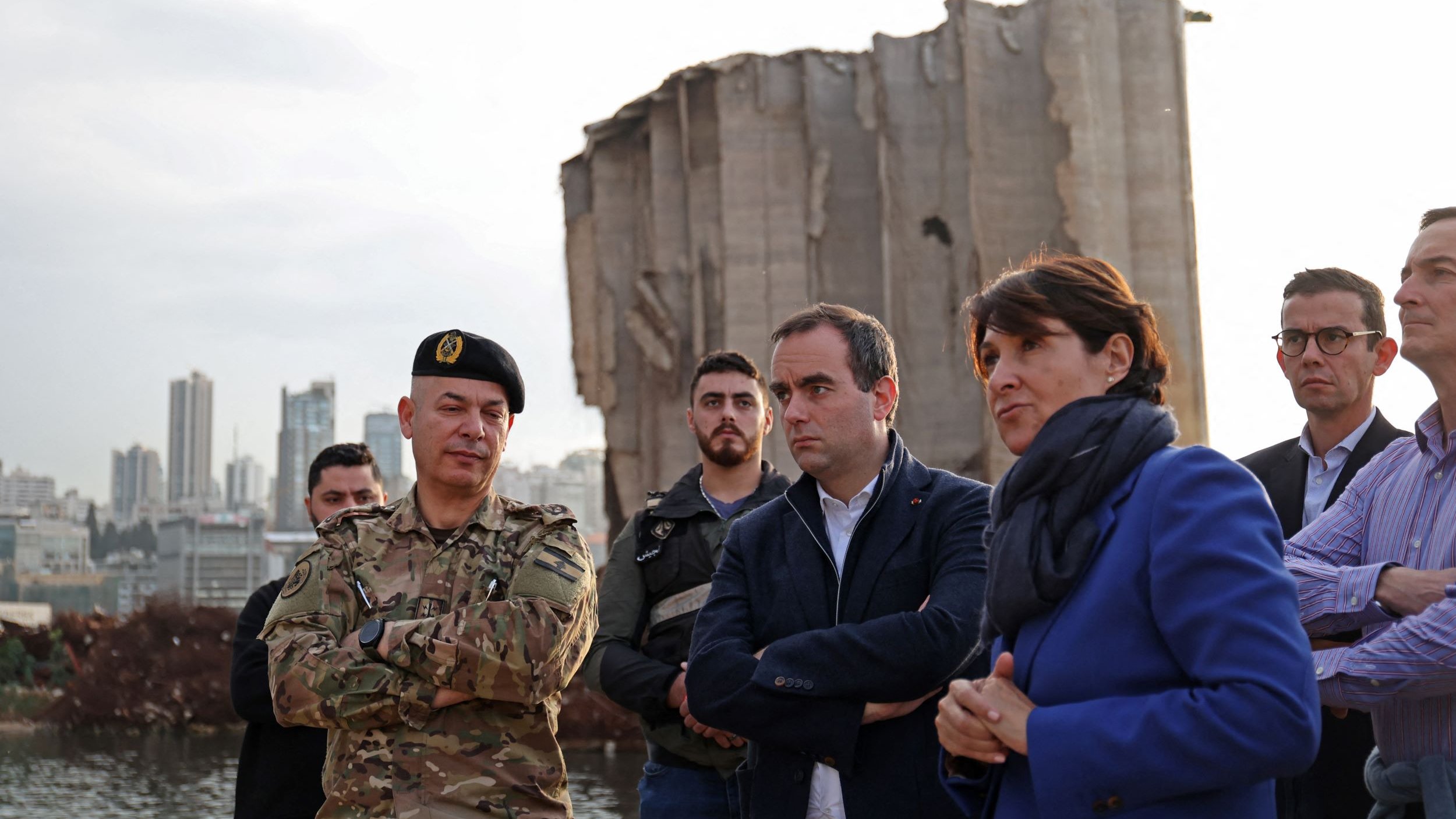 L'image montre plusieurs personnes rassemblées devant une structure endommagée, probablement un bâtiment ou un silo, avec un paysage urbain en arrière-plan. Parmi eux, on distingue un militaire en uniforme, entouré d'autres individus, dont des hommes et des femmes, portant des vêtements civils. L'ambiance semble sérieuse, comme si une discussion ou une évaluation de la situation était en cours. La lumière du jour diminue, suggérant que c'est en fin d'après-midi ou en début de soirée.
