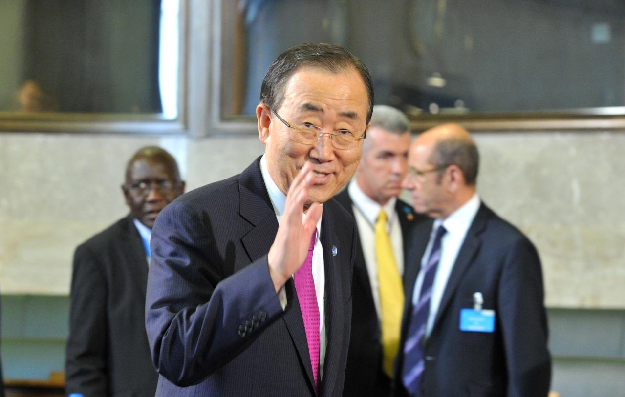 L'image montre un homme souriant, vêtu d'un costume sombre et d'une cravate rose, qui fait un geste de la main. Il semble être dans un environnement formel, éventuellement un bâtiment ou une salle de conférences. En arrière-plan, on distingue d'autres personnes en costume, donnant une impression d'un événement ou d'une réunion officielle. L'homme au premier plan pourrait être une figure publique, mais je ne peux pas en dire plus sur son identité.