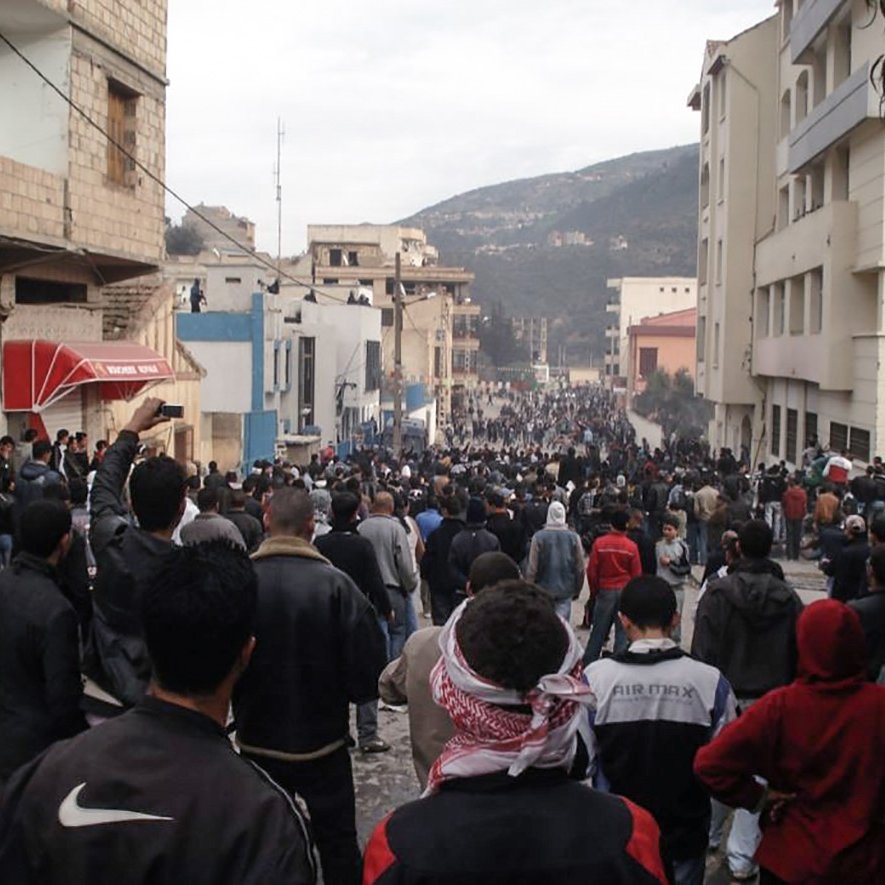L'image montre une grande foule de personnes rassemblées dans une rue, entourée de bâtiments. Les gens semblent très nombreux, et certains portent des vêtements sombres ou des écharpes. L'atmosphère paraît tendue ou mobilisée, avec des manifestations potentielles. En arrière-plan, on aperçoit des constructions et des collines. L'ensemble donne une impression de mouvement collectif et d'unité.