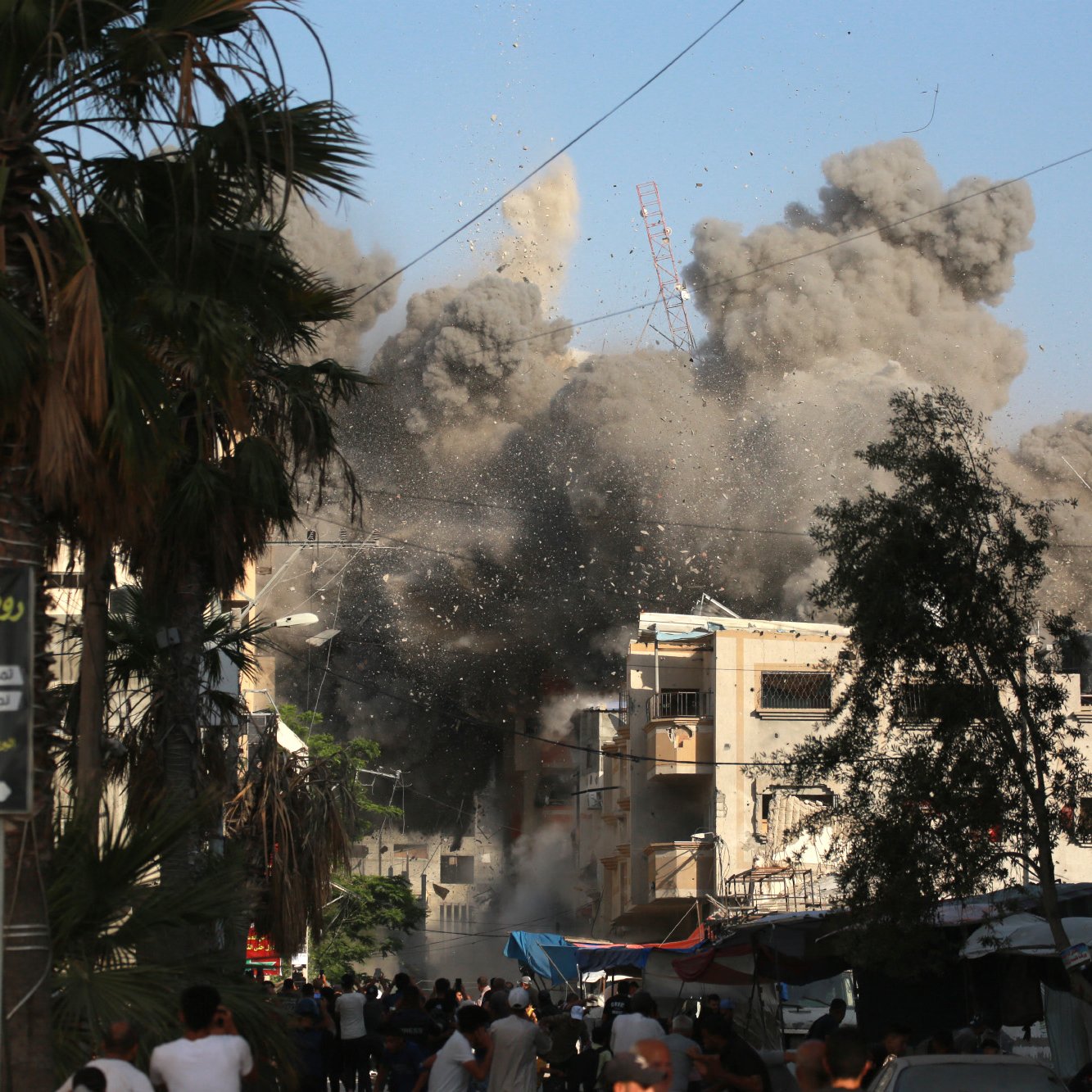 L'image montre une scène chaotique en milieu urbain, où des explosions entraînent un nuage de poussière et de débris. Des bâtiments semblent être endommagés, créant un environnement de destruction. On peut voir des gens dans la rue, réagissant à la situation, ainsi que des palmiers qui ajoutent une touche de verdure au paysage. Le ciel est clair, en contraste avec la fumée épaisse et sombre qui s'élève des ruines.