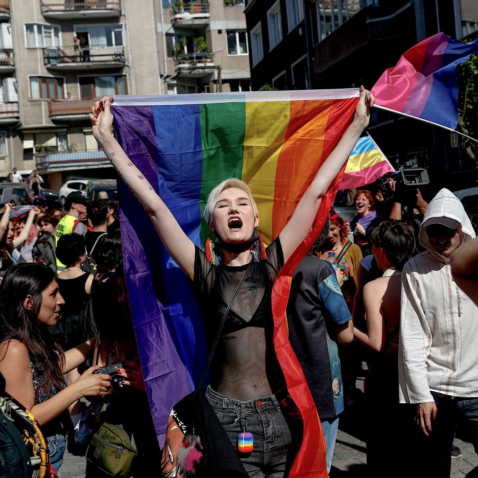 الصورة تظهر مجموعة من الأشخاص في تظاهرة للاحتفال بالفخر والمساواة. في المقدمة، هناك شخص يحمل علم قوس قزح، وهو رمز لحركة حقوق LGBTQ+. يبدو الشخص متحمسًا ويرتدي ملابس مميزة. الخلفية تحتوي على حشود من الناس وبعض الأعلام والمباني، مما يعكس أجواء الاحتفال والتعبير عن الهوية. التجمع يبدو مفعمًا بالحيوية والطاقة.