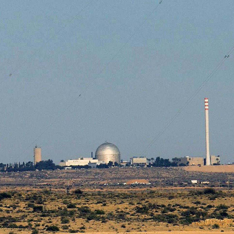 L'image montre un paysage désertique avec des bâtiments en arrière-plan. Au centre, on peut voir un dôme argenté, probablement un réacteur ou une installation scientifique. À côté, il y a une cheminée de grande taille et d'autres structures de différentes hauteurs, suggérant qu'il s'agit d'une installation industrielle ou scientifique. Le ciel est clair et il n'y a pas de nuages. Le terrain autour est aride, avec peu de végétation.