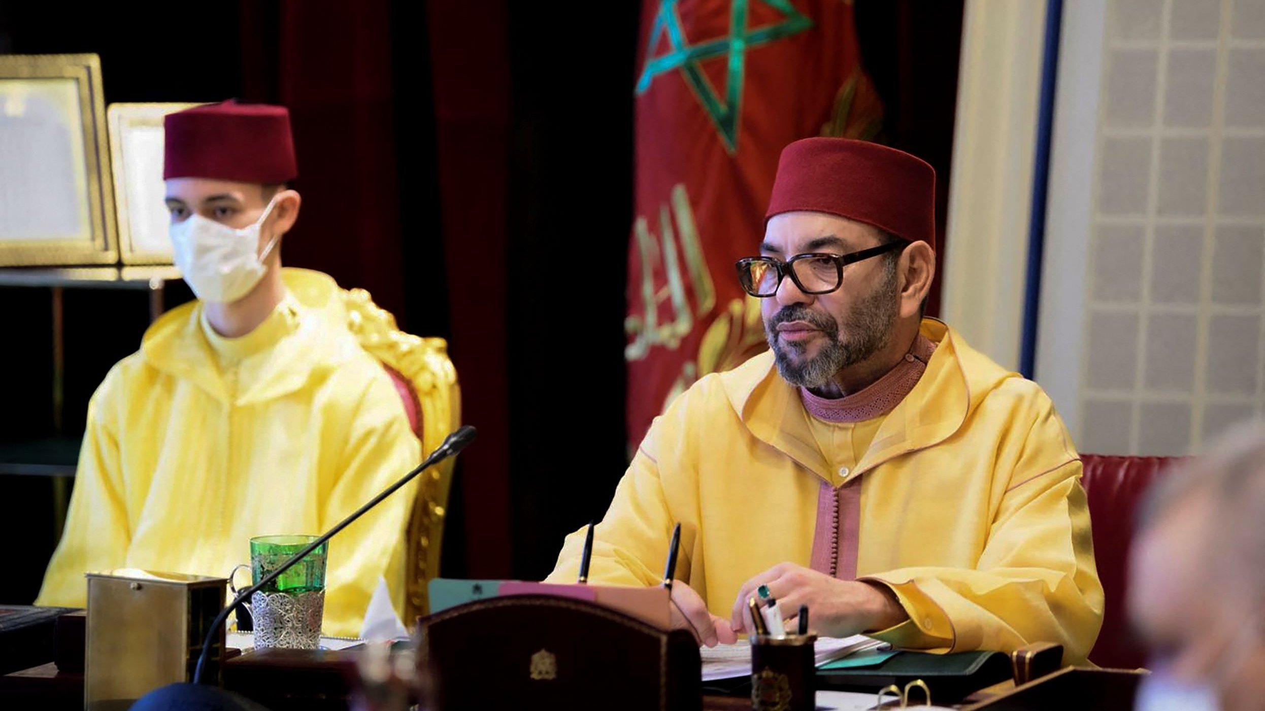 L'immagine mostra un incontro ufficiale in corso. A sinistra, un giovane indossa un abito giallo e una mascherina, mentre a destra c'è un uomo con un berretto rosso e un abbigliamento tradizionale marocchino di colore giallo. L'uomo a destra sembra concentrato, forse mentre prende appunti o partecipa a una discussione. Sullo sfondo si intravedono elementi decorativi di un ambiente formale.