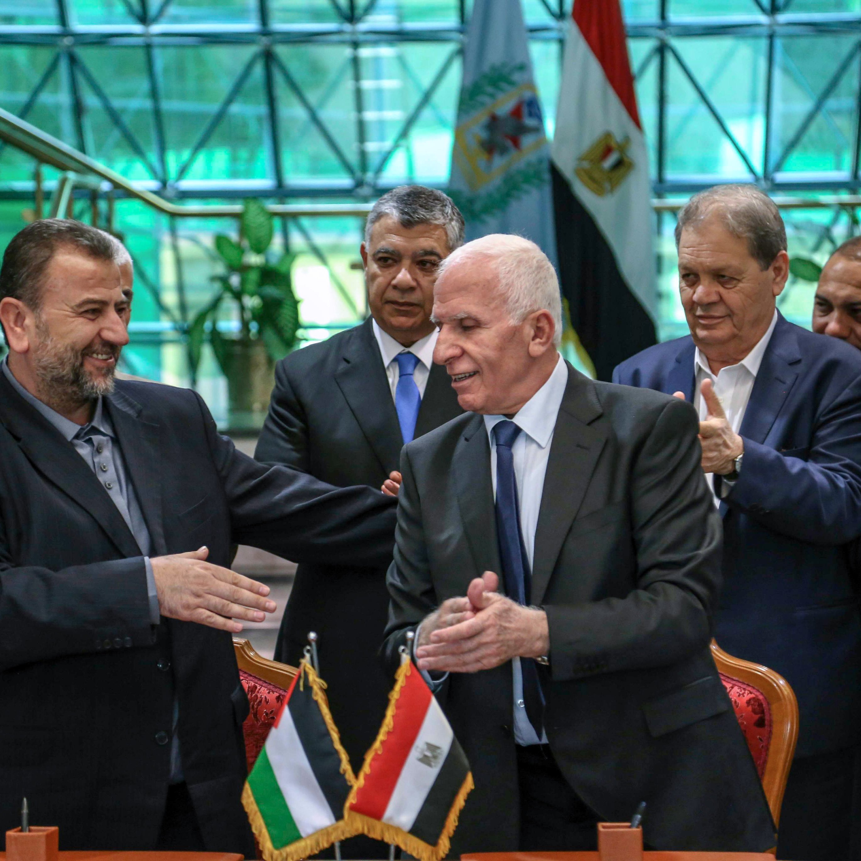 The image shows a formal gathering where two men are shaking hands, likely after a significant agreement or event. They are standing at a table with flags of Palestine and Egypt displayed in front of them. Behind them, a group of men appears to be applauding or showing support. The setting looks official, possibly within a government building, with a modern architectural backdrop. The atmosphere seems positive, indicating a moment of cooperation or partnership.