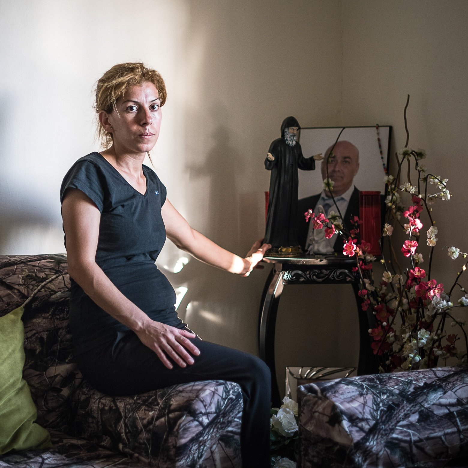 L'image montre une femme assise sur un canapé, portant une tenue noire. Elle est positionnée dans une pièce au mur neutre, avec un éclairage doux. À côté d'elle, il y a un petit meuble sur lequel se trouvent des objets décoratifs, ainsi qu'une photo encadrée d'un homme. Le décor du canapé est à motifs camouflage, et quelques fleurs artificielles ajoutent une touche de couleur à l'environnement. L'expression de la femme semble sérieuse, reflétant peut-être une émotion ou une réflexion profonde.