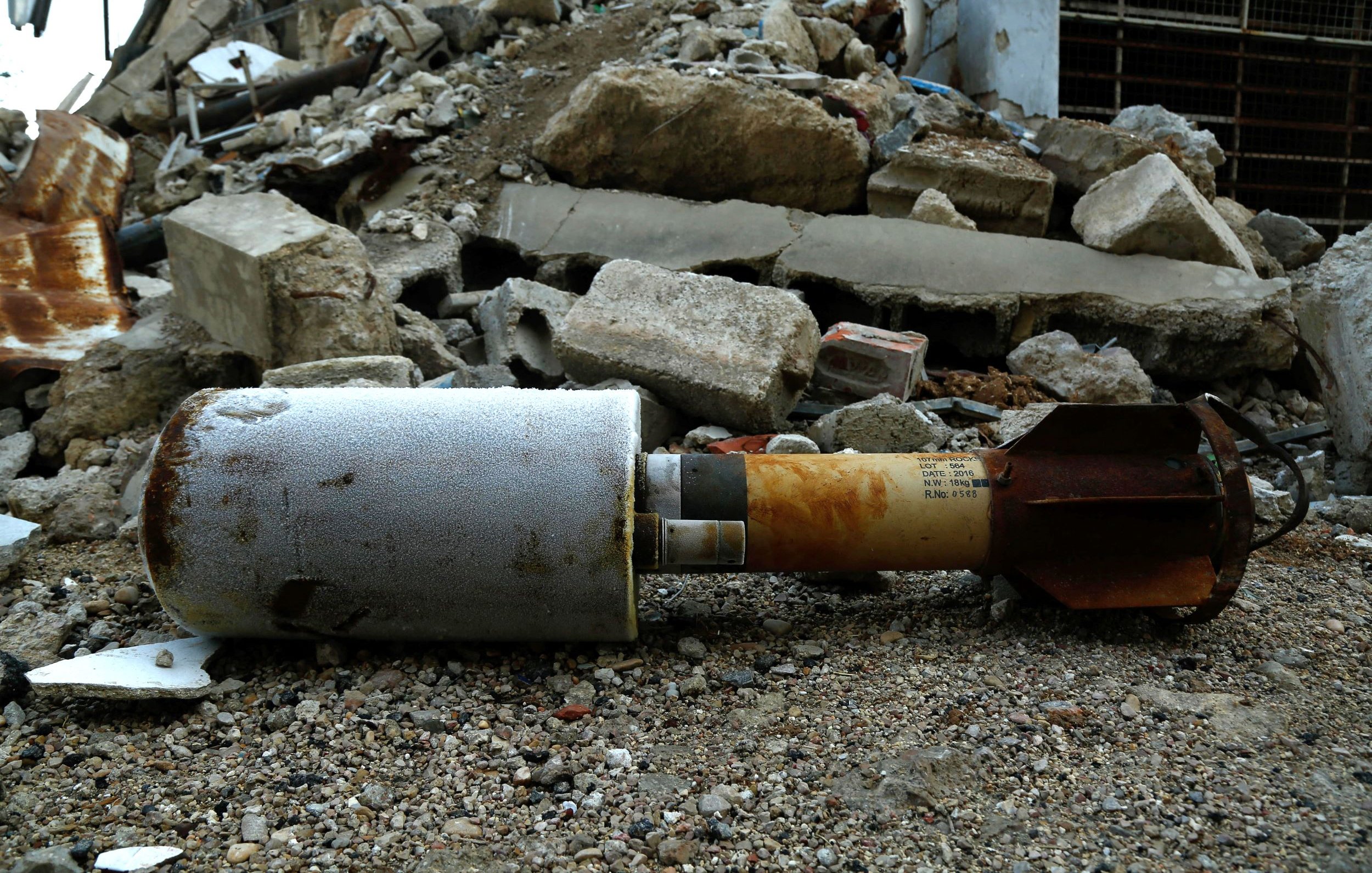 La imagen muestra un artefacto explosivo que yace en el suelo entre escombros de una construcción destruida. El objeto está parcialmente cubierto de polvo y parece haber estado en el lugar durante algún tiempo, lo que sugiere que puede estar relacionado con un conflicto o un evento violento. Los escombros a su alrededor incluyen trozos de concreto y metal, lo que refuerza la impresión de un entorno devastado.
