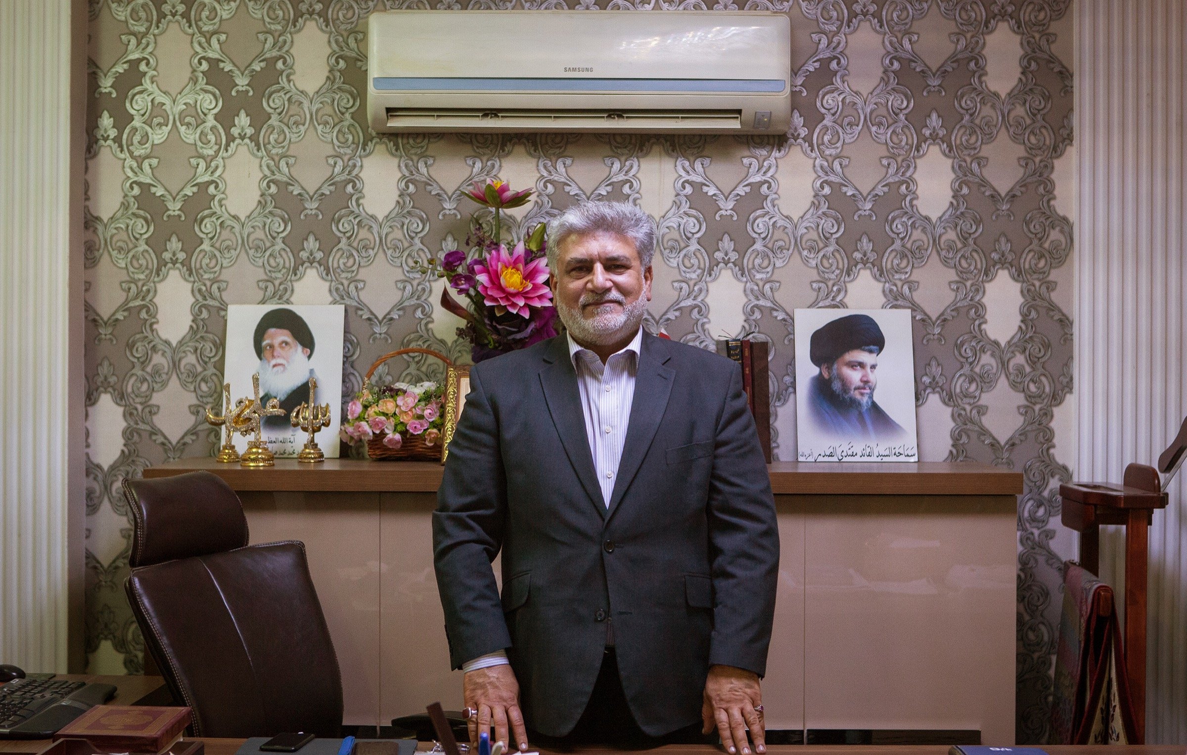 The image shows a man standing in an office setting. He is dressed in a formal suit and has a friendly expression. Behind him, there are two portraits displayed on the wall, likely of influential figures. The office has decorative elements, including a floral arrangement and a neatly organized desk with various items. The overall atmosphere conveys professionalism and authority.