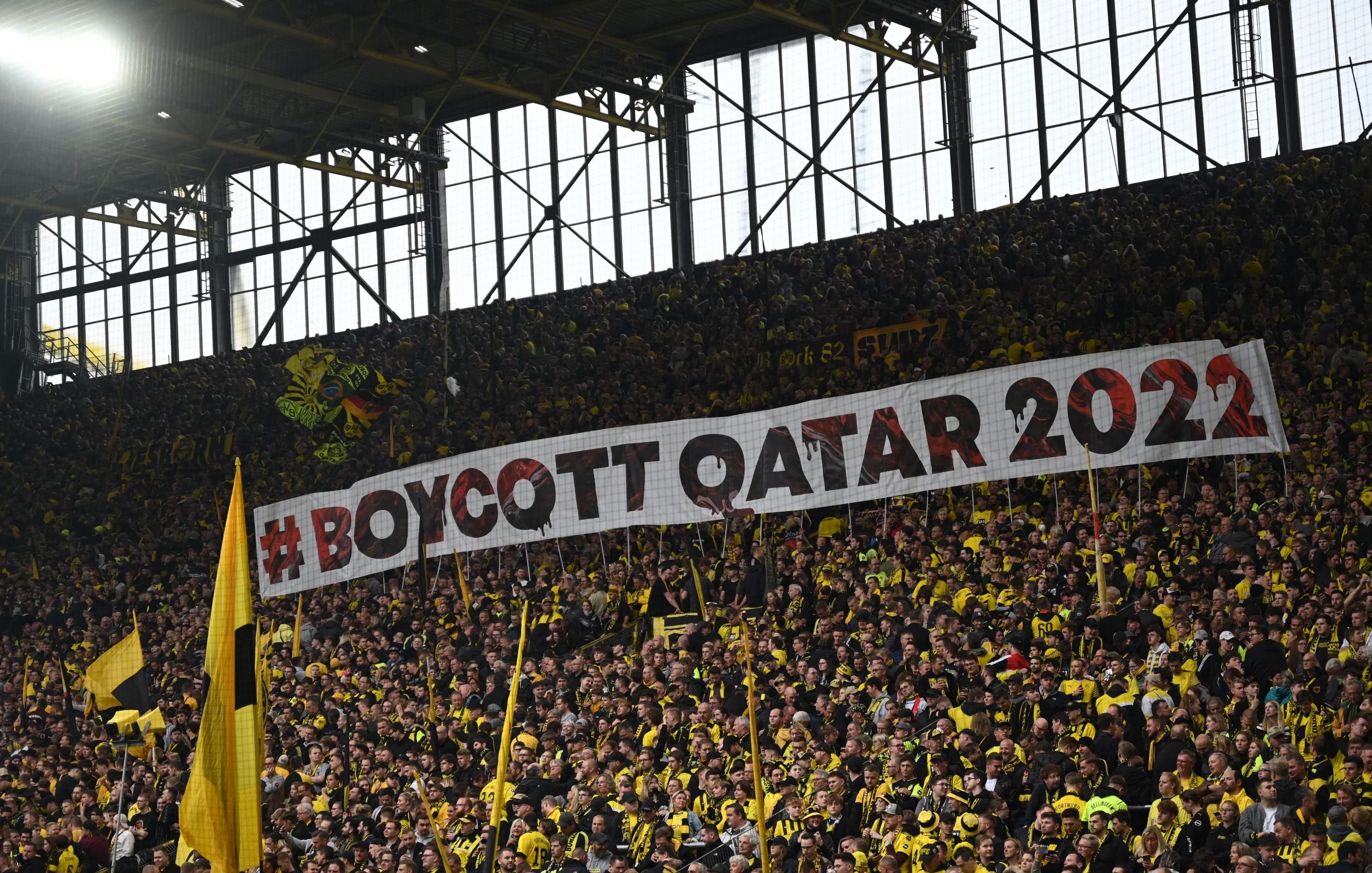 L'image montre un grand groupe de supporters dans un stade, portant des vêtements jaunes et noirs. Ils tiennent une immense banderole sur laquelle est inscrit "#BOYCOTT QATAR 2022". L'atmosphère semble dynamique et engagée, avec un fort sentiment de solidarité parmi les fans. Les tribunes sont pleines, créant une ambiance électrisante.