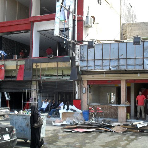 L'image montre un environnement urbain avec des bâtiments partiellement en ruines. On peut voir des façades de bâtiments endommagées, des débris éparpillés au sol et des personnes dans les environs, certaines visiblement en train de travailler sur la reconstruction. Il y a également des voitures garées à proximité. L'atmosphère semble être celle d'une zone en cours de rénovation ou de nettoyage après des dommages.