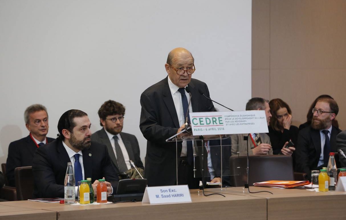 The image depicts a formal conference setting. A middle-aged man, presumably a speaker, is standing at a podium with a sign that reads "CEDRE" and indicates the location and date of the event. He appears to be addressing an audience that includes several men seated behind him, some of whom are engaged in listening or taking notes. The environment has a professional tone, with tables set up and various drinks and papers visible. The atmosphere suggests a significant discussion or presentation, likely focused on economic or policy-related matters.
