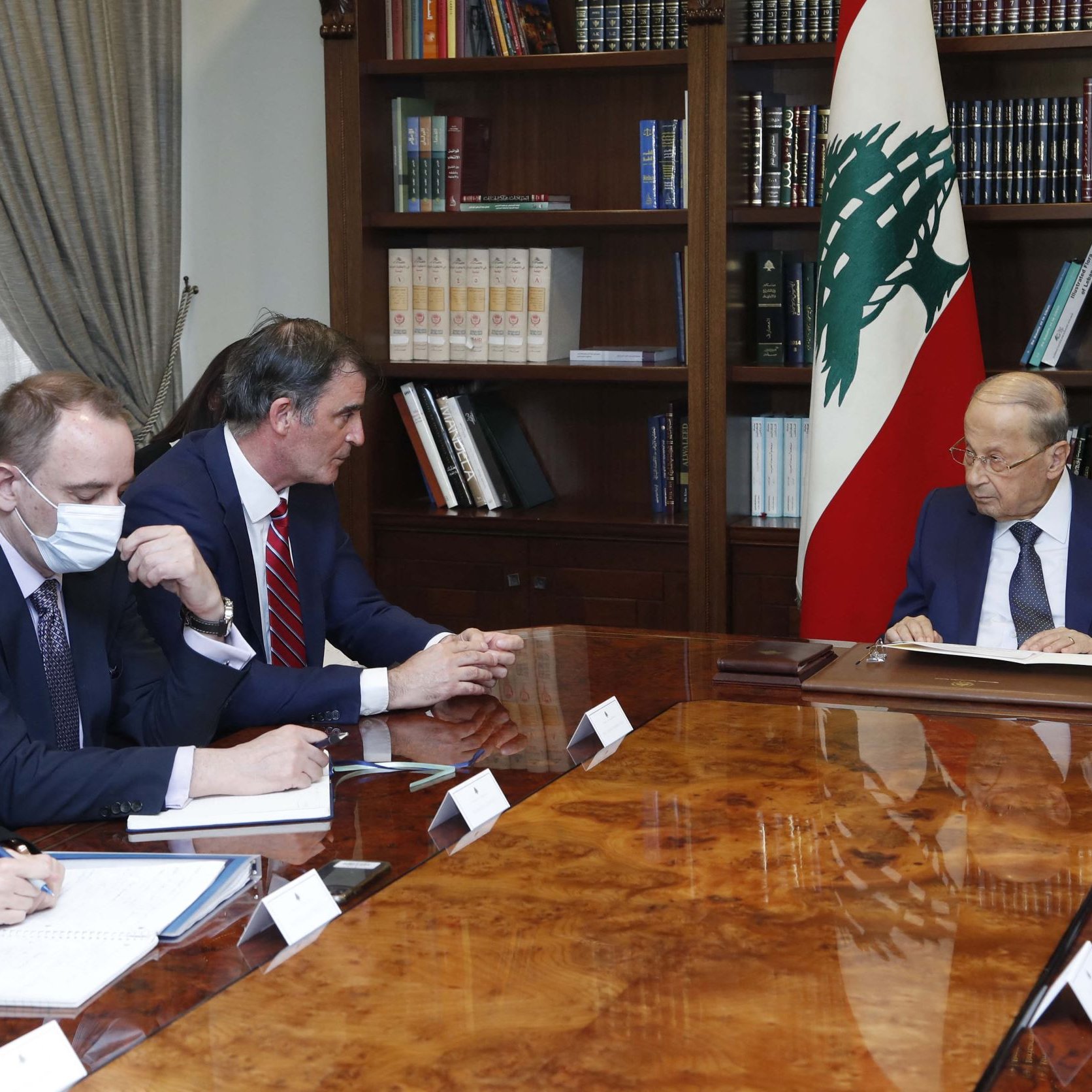 L'image montre une réunion dans un bureau. Un homme, probablement un leader politique, est assis à droite, face à une table en bois. Il semble concentré sur des documents. À sa gauche, il y a trois personnes, vraisemblablement des conseillers ou des diplomates, qui prennent des notes et écoutent attentivement. Tous portent des masques. En arrière-plan, on voit une bibliothèque remplie de livres et le drapeau du Liban. L'atmosphère semble formelle et sérieuse.