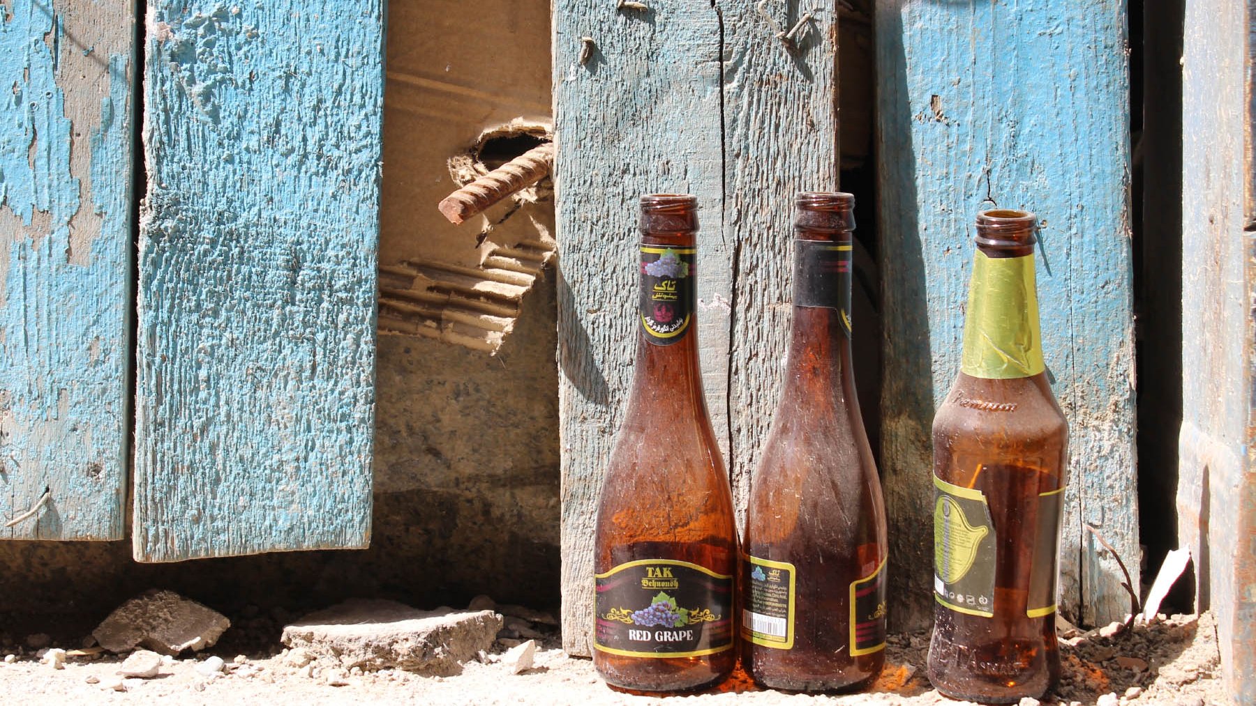 L'image montre trois bouteilles en verre vides, posées sur un sol poussiéreux. Elles sont de formes et de tailles différentes, et montrent des traces de saleté et d'usure. En arrière-plan, on aperçoit des planches de bois bleuies, dont certaines semblent endommagées, avec un trou visible. L'ensemble dégage une impression de désordre et de négligence, suggérant un espace abandonné ou en déclin.