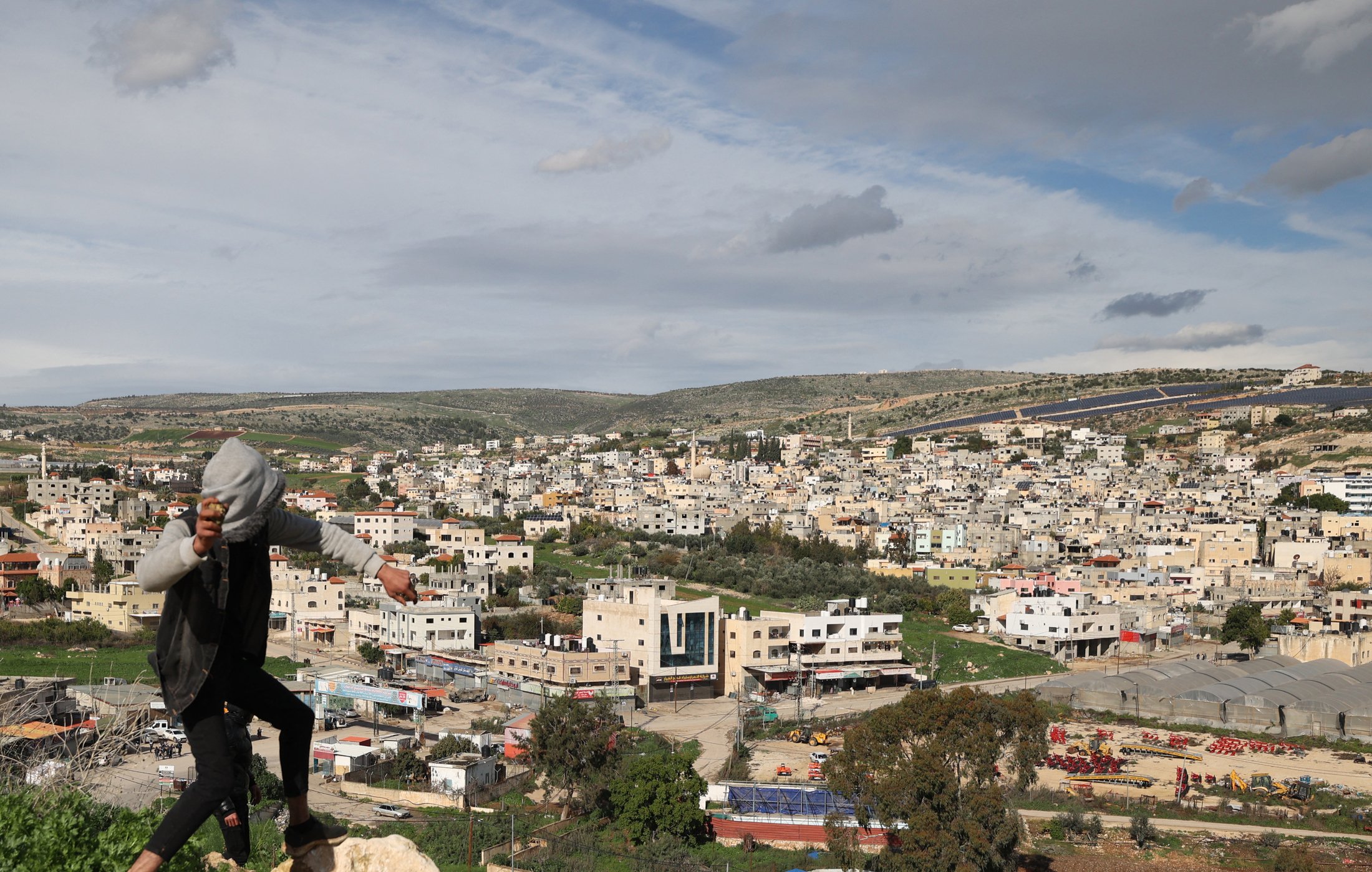 در این تصویر، فردی در حال بالا رفتن یا حرکت در مکانی کوهستانی دیده می‌شود. او لباس‌های غیررسمی به تن دارد و در پس‌زمینه، یک شهر بزرگ با ساختمان‌های مختلف و معماری متنوع دیده می‌شود. آسمان ابری است و مناظر طبیعی اطراف، مانند تپه‌ها و درختان، نیز در تصویر قابل مشاهده‌اند. این صحنه ترکیبی از زندگی شهری و طبیعت را نشان می‌دهد.