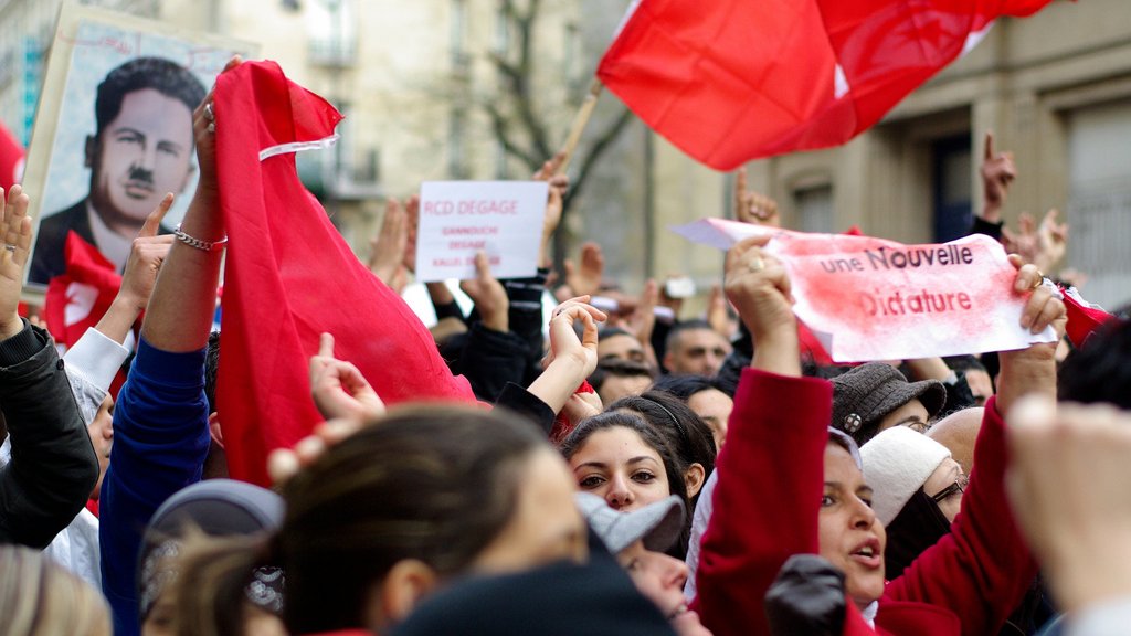 L'image montre une foule de manifestants rassemblés, brandissant des drapeaux rouges et des pancartes. Certains visages sont masqués, tandis que d'autres expriment des émotions intenses, comme la détermination ou la colère. On aperçoit aussi un portrait d'une figure emblématique en arrière-plan. Les slogans sur les pancartes semblent appeler au changement et à la liberté, indiquant un contexte de protestation politique. L'atmosphère est chargée d'énergie collective.