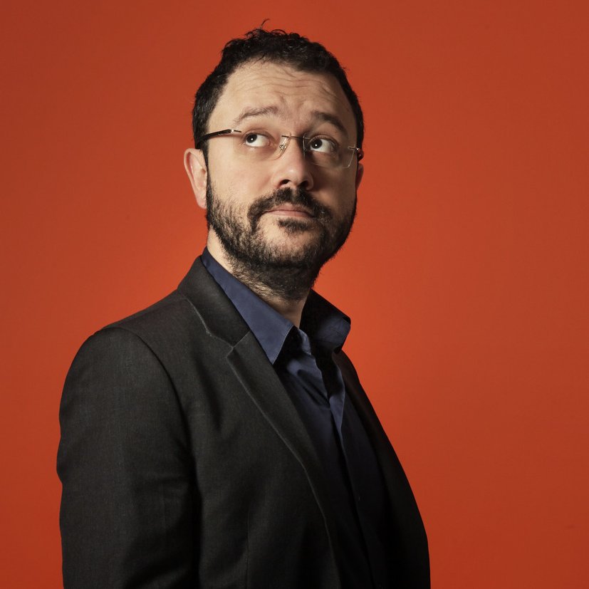 L'image montre un homme vêtu d'un costume noir sur une chemise bleu foncé, regardant légèrement sur le côté. Il a des lunettes, une barbe et un léger sourire. L'arrière-plan est orange, ce qui crée un contraste vif et dynamique avec sa tenue sombre. L'expression de son visage semble pensif ou intrigué.