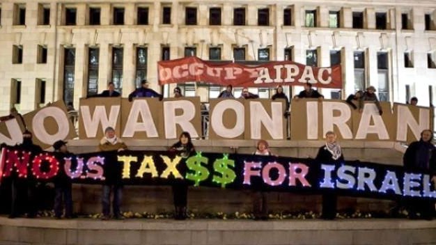 L'image montre un groupe de personnes réunies autour d'une grande pancarte ou d'un affichage, exprimant des revendications politiques. Ils tiennent des panneaux où l'on peut lire "NO WAR ON IRAN" et "NO US TAX$$ FOR ISRAEL". L'ambiance semble être celle d'une manifestation ou d'un rassemblement en faveur de la paix et critiquant l'utilisation des fonds publics. L'arrière-plan comprend un bâtiment, suggérant que l'action a lieu dans un espace public.