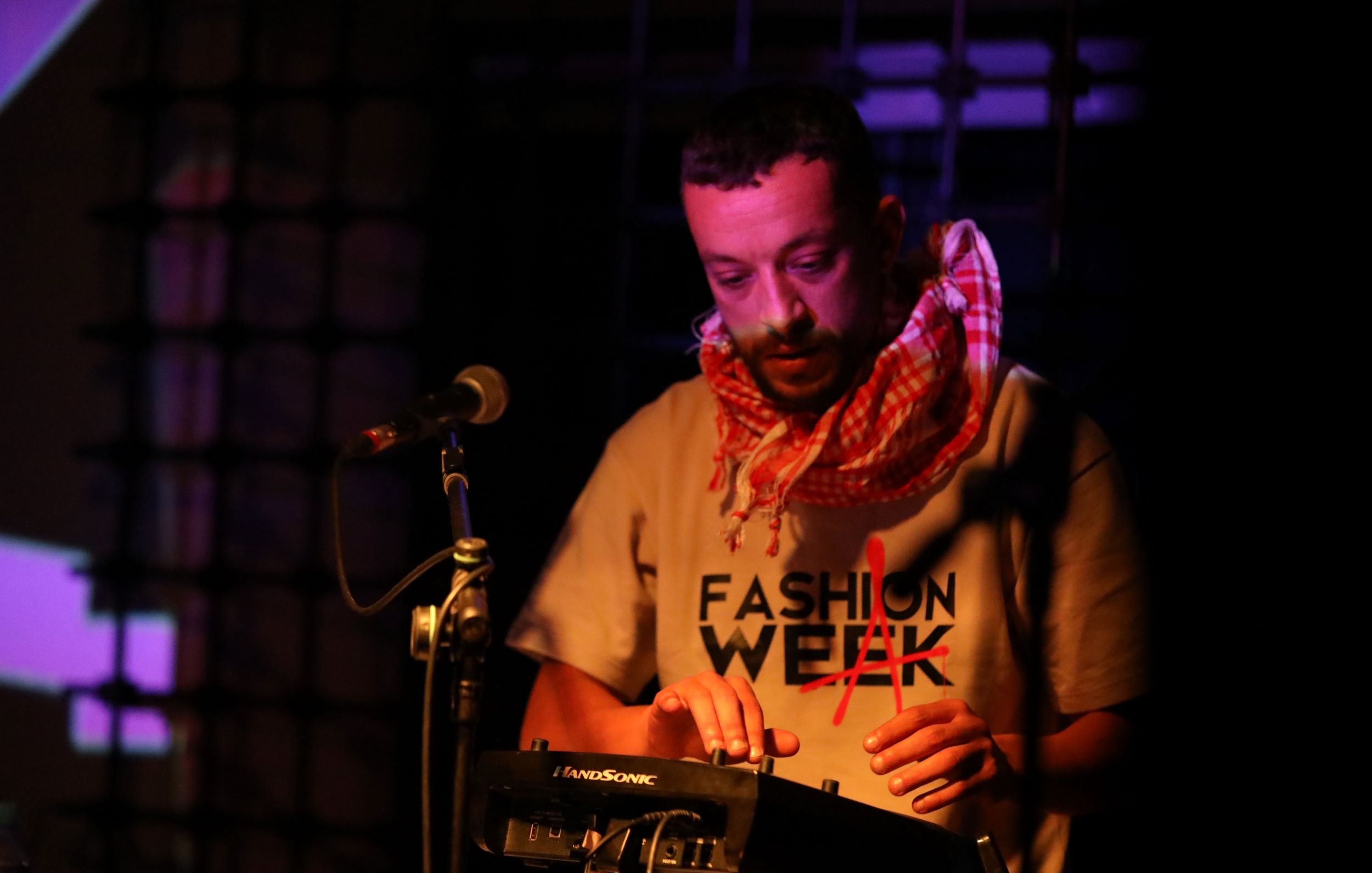L'immagine mostra un musicista che sta esibendosi su un palco. Indossa una maglietta con la scritta "FASHION WEEK" e una sciarpa a quadri. È concentrato mentre suona un dispositivo elettronico, probabilmente un sintetizzatore o una drum machine. Il palco è illuminato da luci colorate che creano un'atmosfera vivace e coinvolgente. Dietro di lui, si intravedono proiezioni visive che aggiungono dinamismo alla performance.