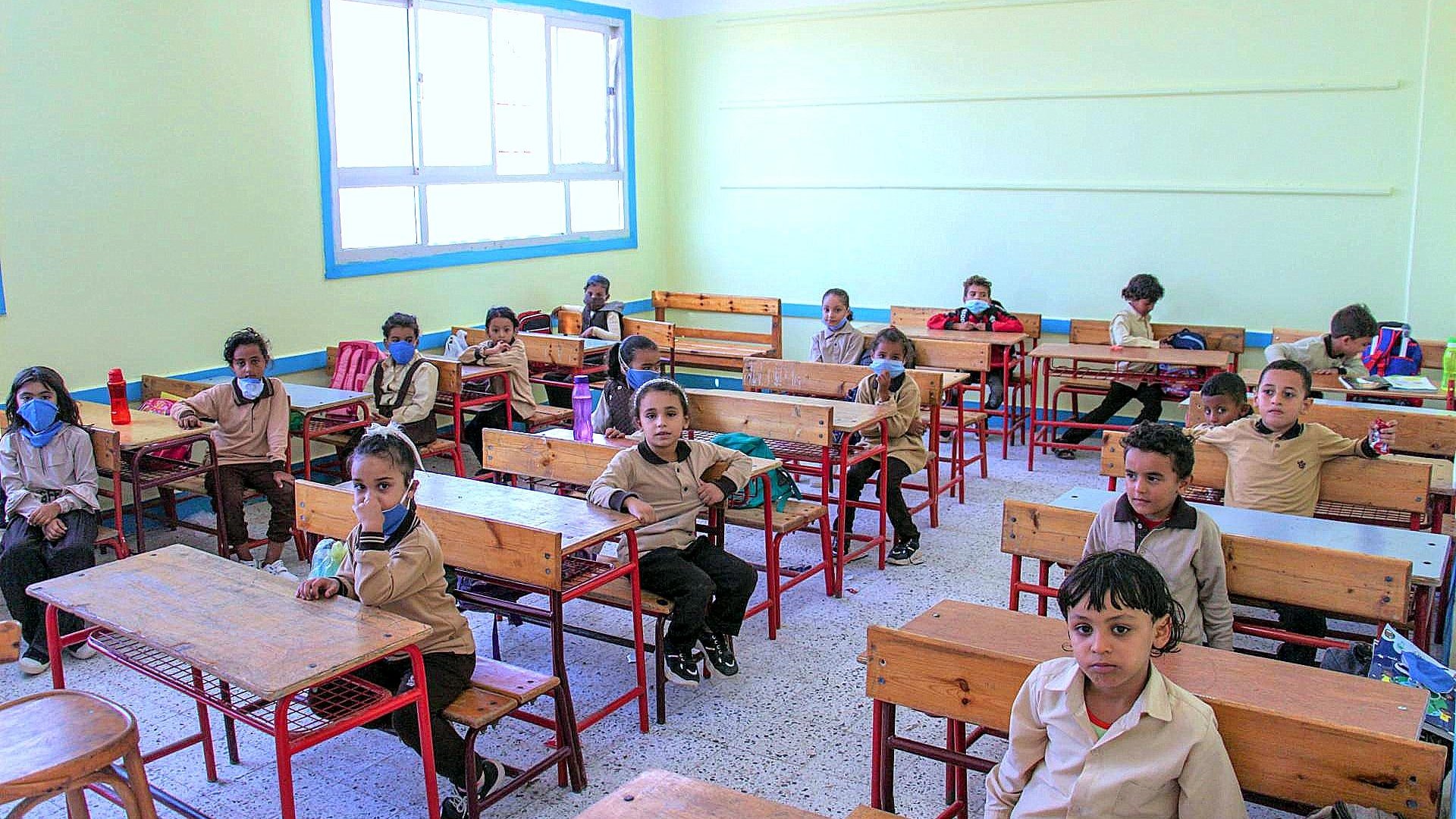 الصورة تُظهر فصلاً دراسياً يحتوي على عدد من الأطفال في مقاعدهم. الغرفة مضاءة جيداً بألوان فاتحة، الجدران مطلية بلون أخضر فاتح. الأطفال يرتدون زي المدرسة ويبدو عليهم الانتباه. بعضهم يرتدي كمامات. الطاولات والكراسي مصنوعة من الخشب. الغرفة منظمة ومرتبة وتحتوي على نوافذ كبيرة تسمح بدخول الضوء الطبيعي.