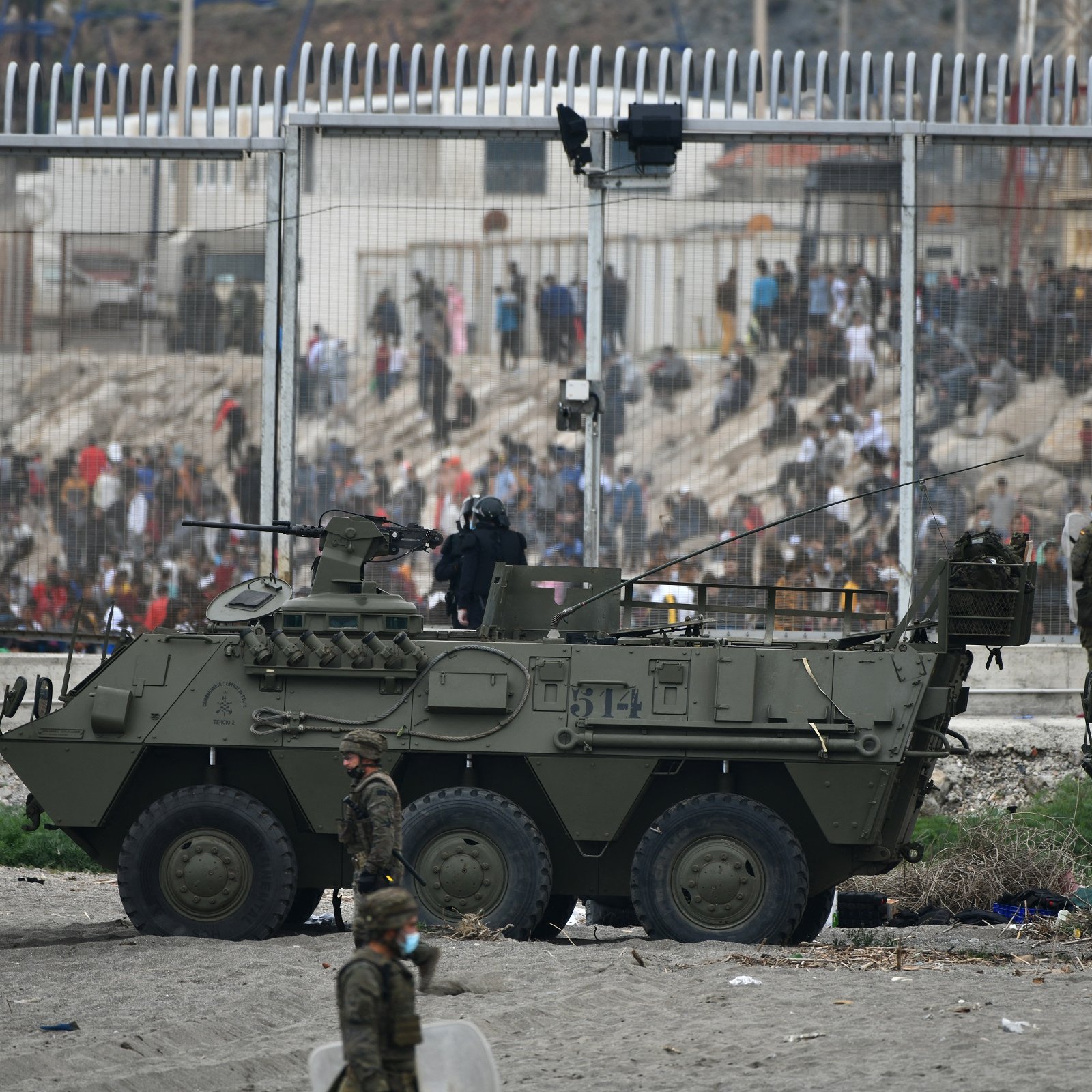 La imagen muestra un contexto de tensión, probablemente en una zona de conflicto o crisis. Se pueden ver vehículos militares blindados en la parte frontal, con soldados a bordo y personal militar en el entorno. Al fondo, hay una multitud de personas que parecen estar frente a una valla alta, indicando una separación entre diferentes grupos. El ambiente es de intensa actividad, posiblemente con indicios de enfrentamientos o una situación de disturbio. La escena evoca un fuerte sentido de vigilancia y seguridad militar.