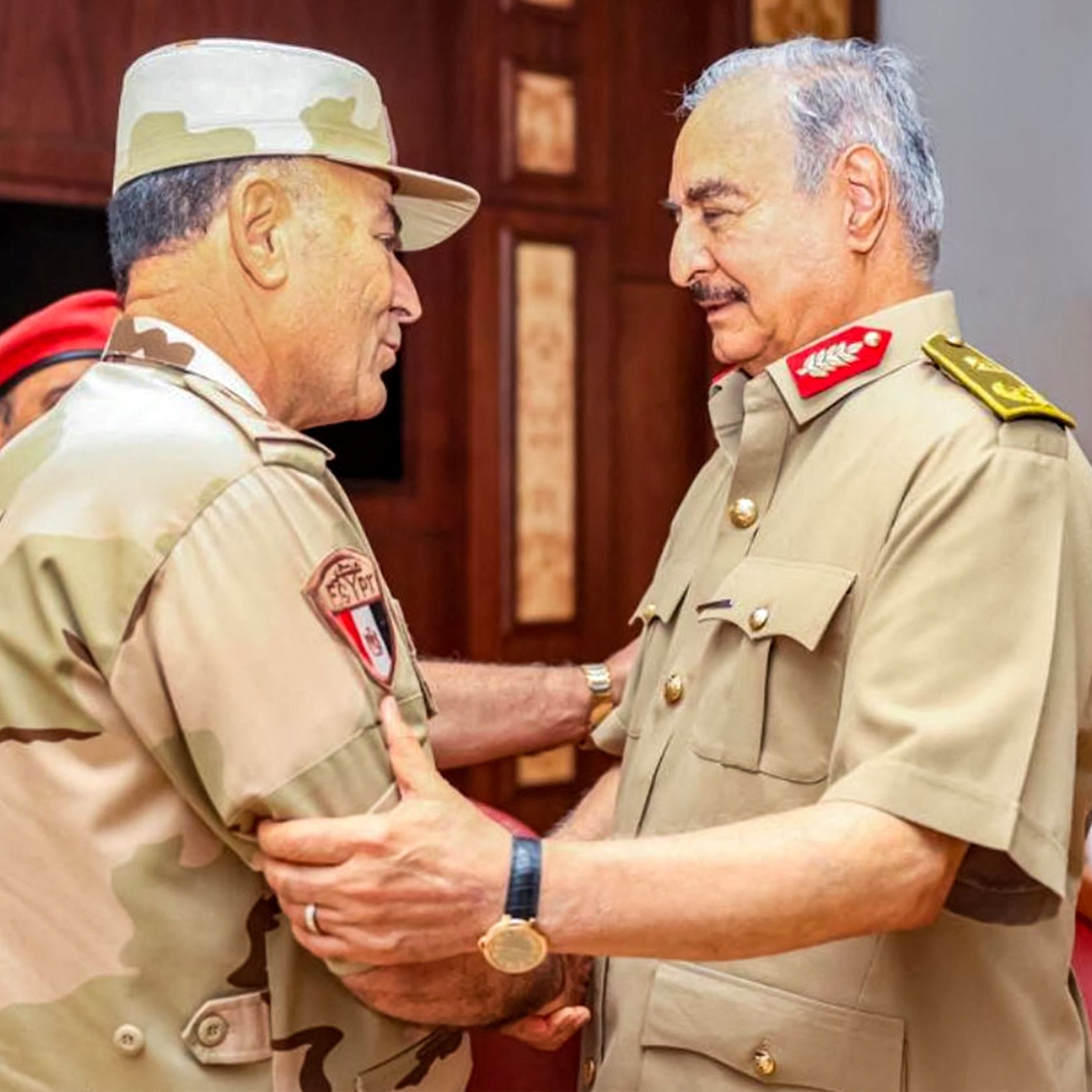 L'image montre deux militaires en uniforme, se tenant face à face et en train de discuter. L'un d'eux porte un chapeau militaire et un uniforme beige avec des motifs camouflage, tandis que l'autre est habillé en uniforme militaire beige et porte des décorations sur son épaule. Ils semblent avoir une conversation amicale et posent dans un environnement intérieur, avec des tables et des chaises visibles en arrière-plan. L'atmosphère semble informelle, suggérant une réunion ou un échange entre officiers.