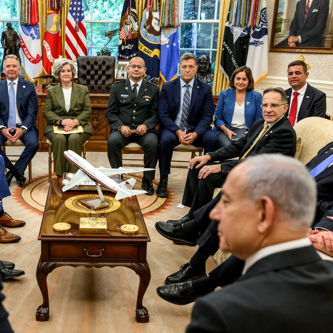 A formal meeting in the Oval Office with various officials and a model airplane on the table.