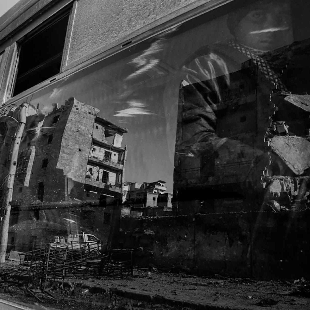 L'image présente une scène en noir et blanc, où un bus est au premier plan. On peut voir le reflet d'un homme, probablement en train de porter un vêtement sombre et une écharpe, à côté d'une architecture en ruine. À travers la vitre du bus, des bâtiments dégradés sont visibles, indiquant des signes de destruction. L'atmosphère semble sombre et tragique, évoquant des thèmes de dévastation et de résilience.