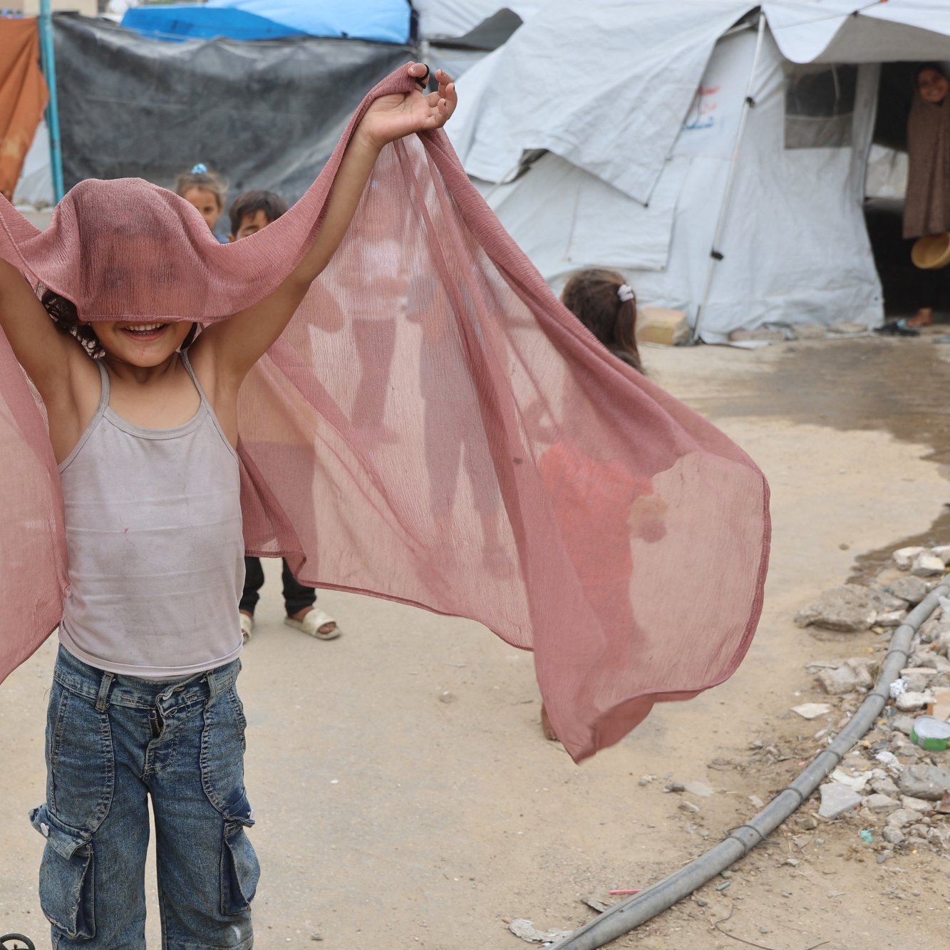 A smiling child holds up sheer fabric, playfully expressing joy in a makeshift camp.