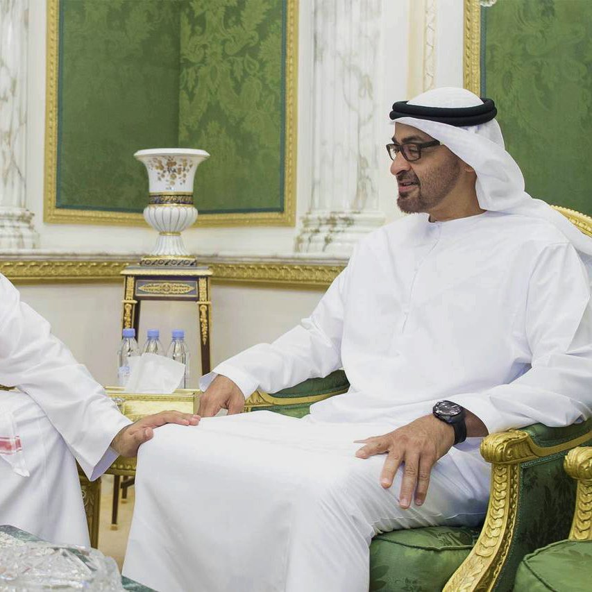 تظهر في الصورة اثنان من الرجال يجلسان معًا في غرفة مزينة بطريقة فاخرة. يرتدي الأول الزي السعودي التقليدي مع الشماغ الأحمر والأبيض، بينما يرتدي الثاني الزي الإماراتي التقليدي. كلاهما يبدو سعيدًا، ويتبادل الحديث في أجواء ودية. الخلفية تتضمن تفاصيل خضراء وذهبية، مما يضيف جوًا من الرقي والفخامة. على الطاولة أمامهم زجاجات ماء، مما يشير إلى اجتماع أو مناسبة اجتماعية.