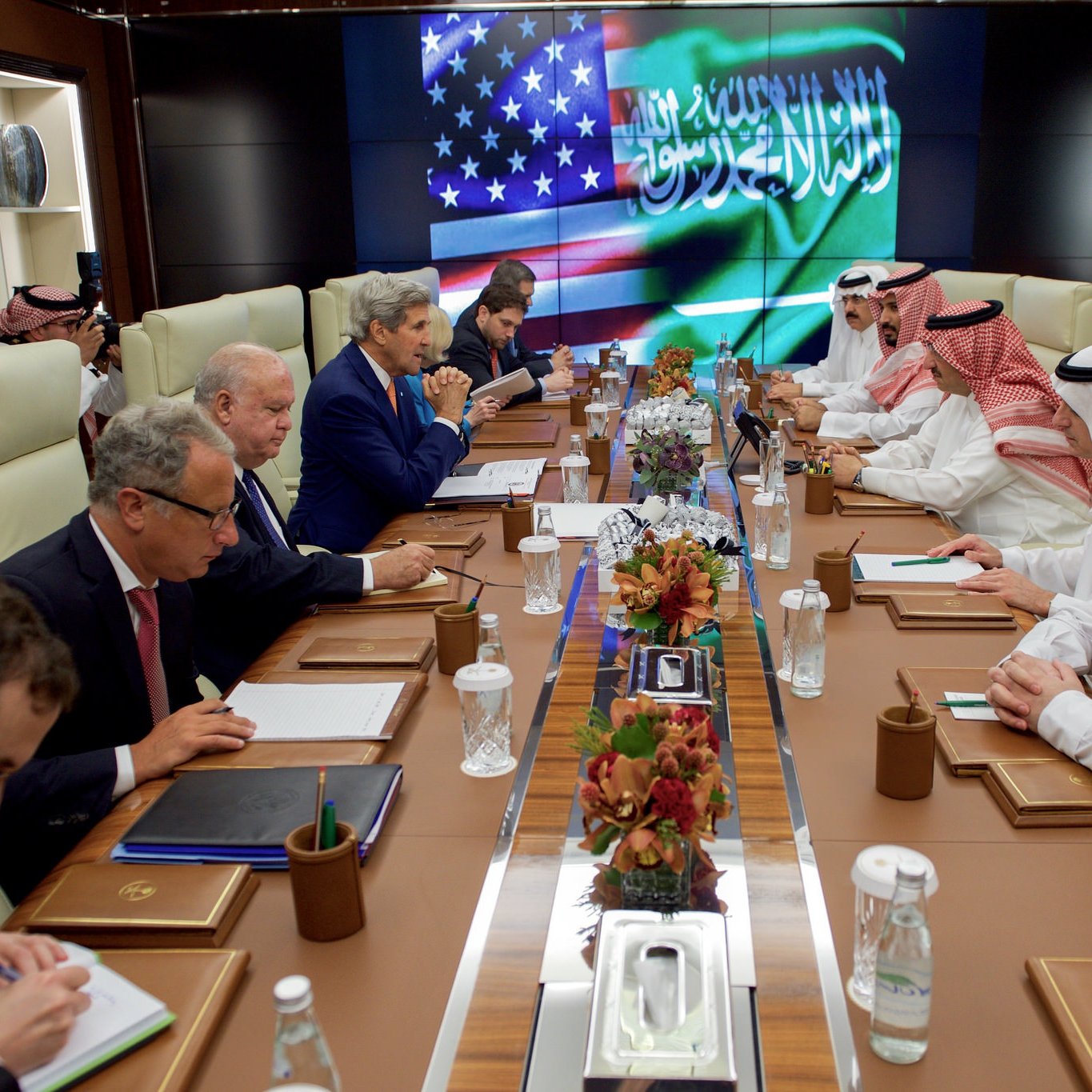 تظهر الصورة اجتماعًا رسميًا يجمع بين مسؤولين سعوديين وأمريكيين. في غرفة تجمعهم، يتواجد ممثلون من كلا الجانبين، حيث يجلس الرجلان في المقدمة على طاولة مستديرة، بينما يقف آخرون بجانبهم. هناك شاشة في الخلف تعرض علمي السعودية والولايات المتحدة، مما يعكس شراكة أو تعاون بين البلدين. يبدو أن الجميع يركز على المناقشات الجارية مع وجود ملاحظات وأدوات على الطاولة.