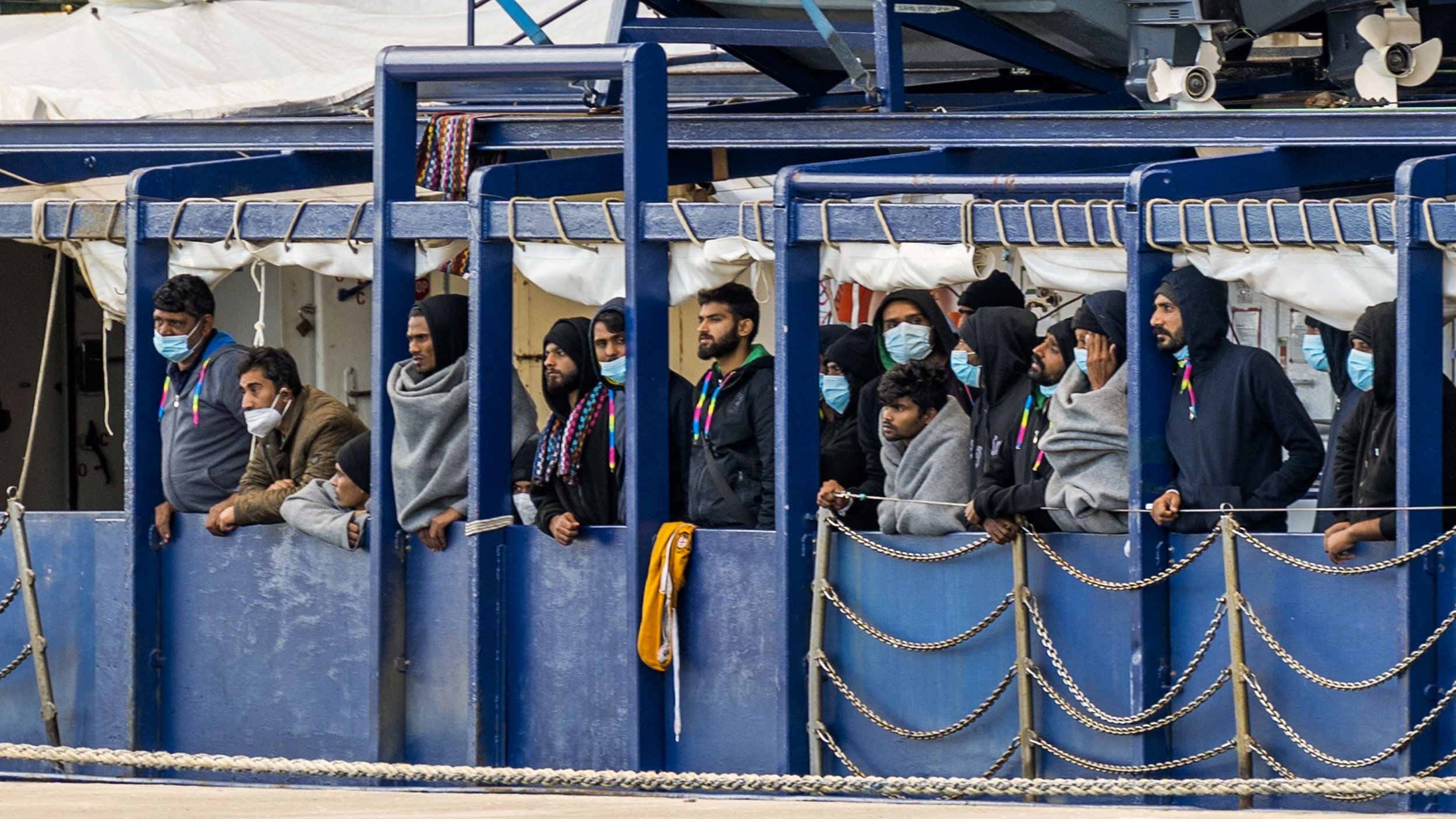 L'image montre un groupe de personnes se tenant sur un bateau. Elles portent des masques et des vêtements semblant chauds, comme des sweats à capuche. Les gens regardent vers l'extérieur, probablement vers la terre ou vers un port. L'environnement semble portuaire, avec des équipements et d'autres structures en arrière-plan. L'atmosphère peut sembler préoccupante ou attendue, reflétant une situation particulière.