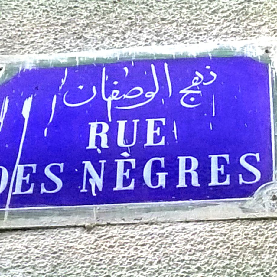 تظهر في الصورة لافتة شارع مكتوبة باللغة الفرنسية والعربية. الخلفية لونها أزرق، وتحتوي على نص باللون الأبيض. النص الفرنسي يقول "RUE DES NÈGRES"، بينما النص العربي يكتب "ذراع الوصفان". يبدو أن اللافتة قديمة بعض الشيء، حيث تظهر عليها آثار الط weather والاتساخات.
