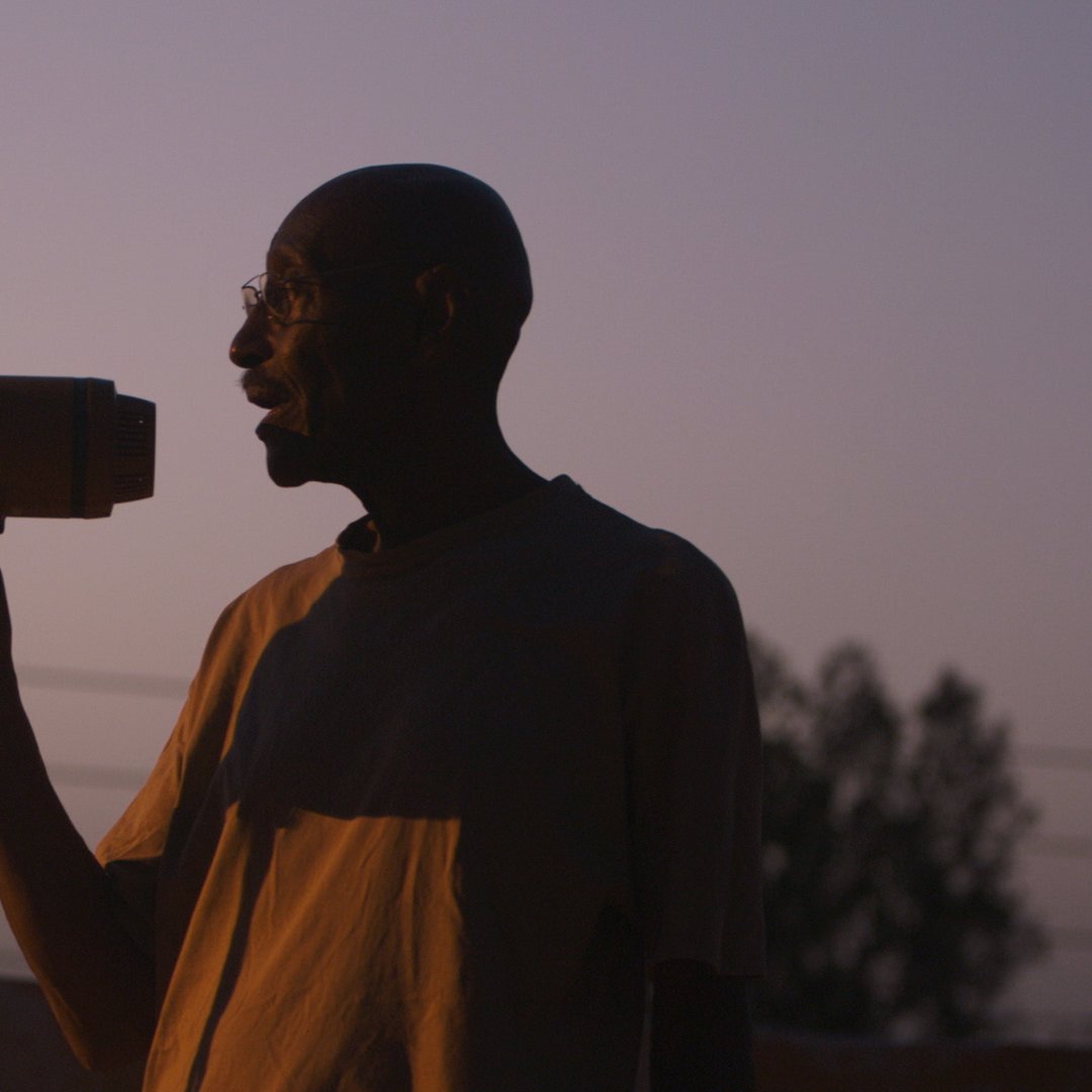 رجل يحمل مكبر صوت، silhouetted أمام غروب الشمس، يبدو وكأنه ينادي بشيء مهم.