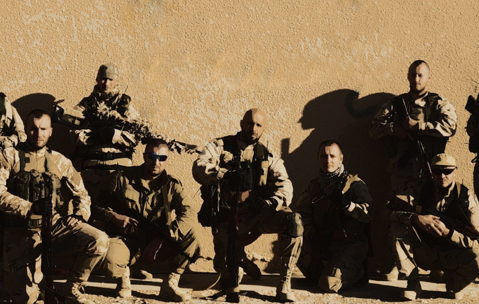 The image appears to show a group of military personnel posing together against a neutral-colored background. They are dressed in tactical gear, including vests and uniforms, and are equipped with firearms. The group is arranged in two rows, with some members kneeling and others standing. The overall tone of the image suggests a camaraderie among the team. The lighting emphasizes their serious expressions and the rugged environment.