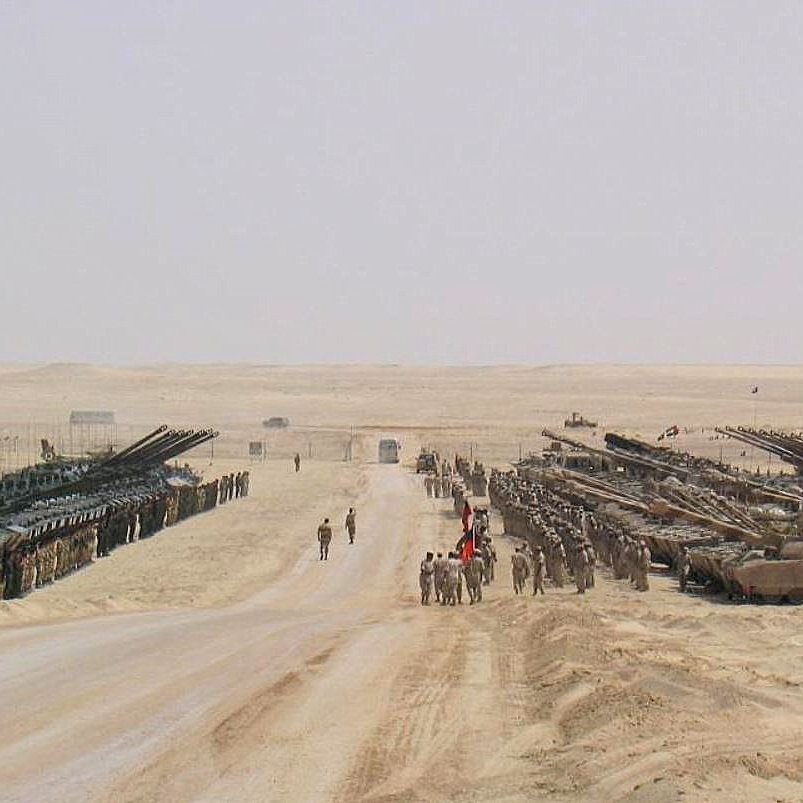 The image depicts a vast desert landscape, characterized by a flat, sandy terrain. On either side of a wide dirt road, there are rows of military tanks and armored vehicles arranged in a formation. Several soldiers are visible on the road, possibly coordinating or observing the vehicles. The background features an expansive desert with minimal vegetation, emphasizing the military setup in a stark and barren environment. The scene conveys a strong military presence and readiness.