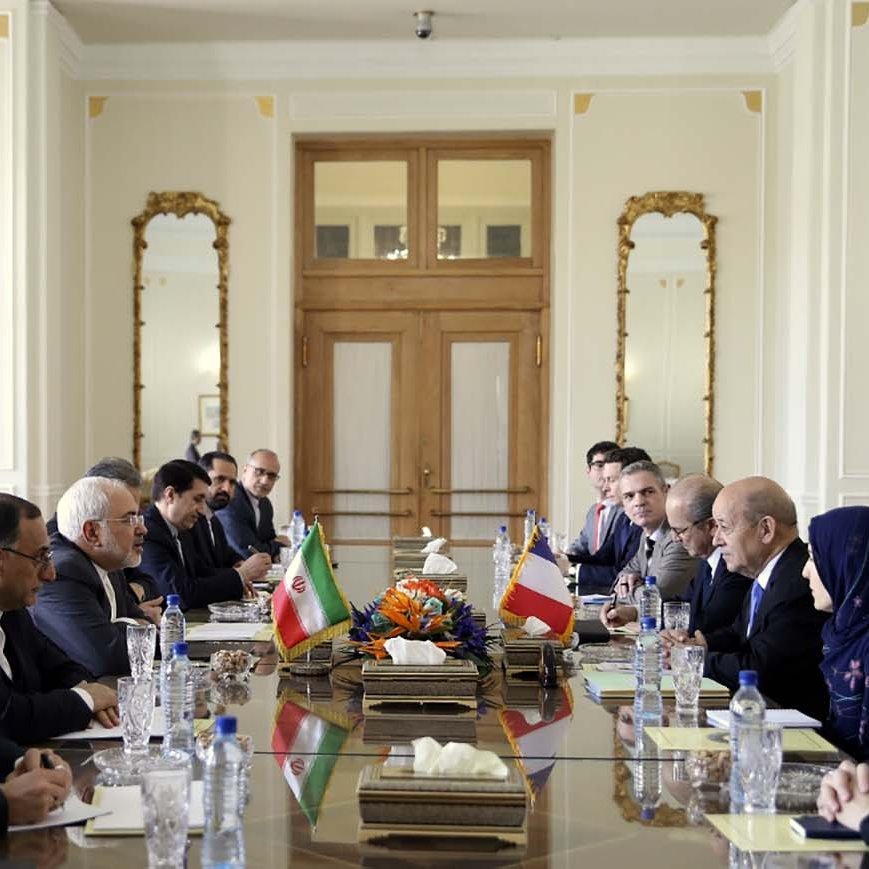 L'image montre une salle de réunion avec une grande table au centre. De chaque côté de la table, plusieurs personnes sont assises, participent à une discussion. Les drapeaux de l'Iran et d'un autre pays sont visibles sur la table. L'environnement est élégant, avec des décorations raffinées et de grandes fenêtres. On peut aussi voir des fleurs au centre de la table. Les participants semblent concentrés et engagés dans la conversation.