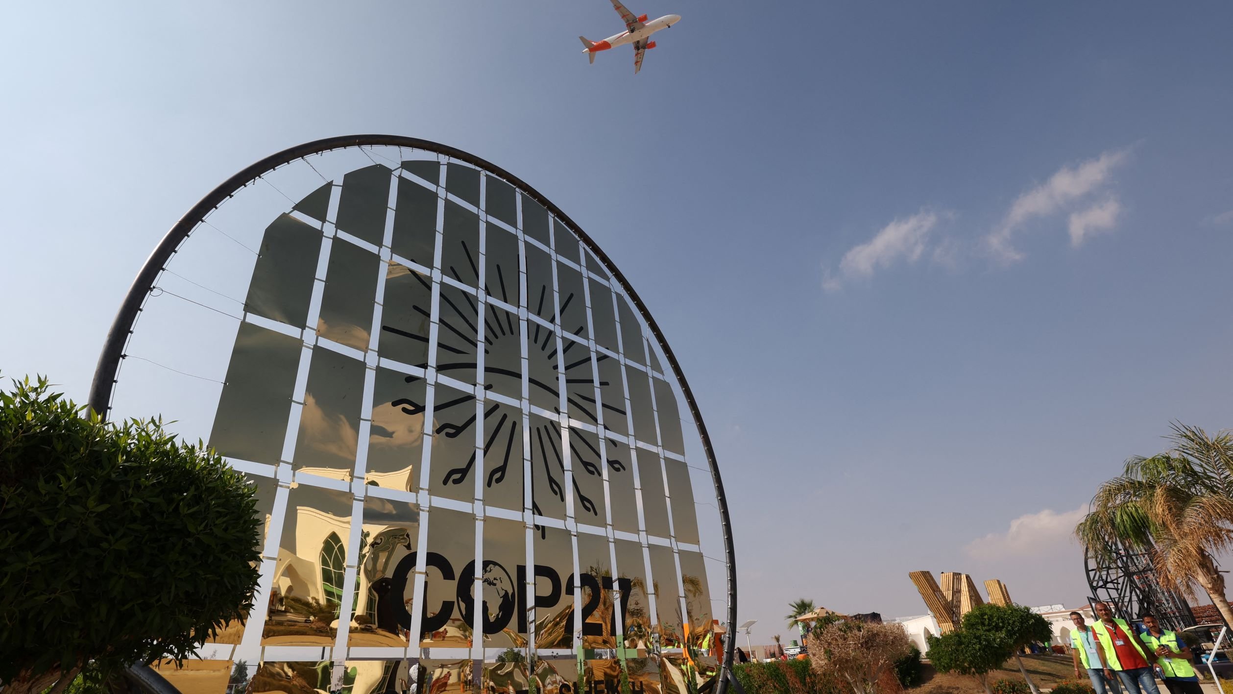 L'image montre un panneau de bienvenue à la COP27, ornée d'un design en miroir qui reflète le ciel. On aperçoit un avion volant au-dessus, suggérant une activité aérienne dans la région. Le paysage présente des éléments végétaux, comme des plantes et des arbres, et des personnes portant des vestes de sécurité sont visibles, indiquant une ambiance de préparation pour l'événement. Le ciel est clair avec quelques nuages.