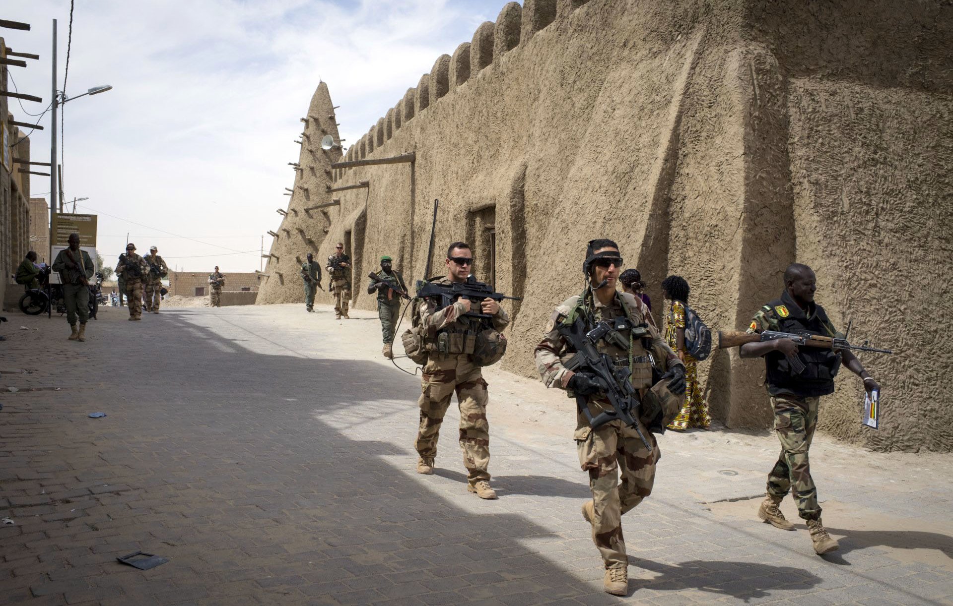 The image depicts a group of soldiers walking through a narrow street in a fortified area. They are dressed in military gear and armed with rifles. The architecture features traditional, mud-brick structures, which create a historical and cultural backdrop. The scene conveys a sense of military presence and activity, with more soldiers seen in the background, indicating a coordinated operation or patrol. The atmosphere appears to be tense and vigilant, typical of a military environment.