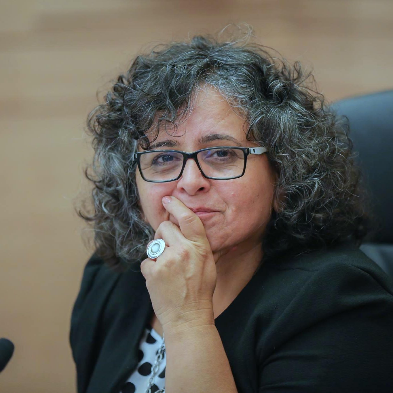 L'image montre une femme avec des cheveux bouclés et un regard réfléchi. Elle porte des lunettes et une veste. Une main est placée sur son menton, ce qui suggère qu'elle est en train de penser ou de considérer une question. L'environnement semble être une salle de réunion ou une assemblée, avec des chaises et une table en arrière-plan.