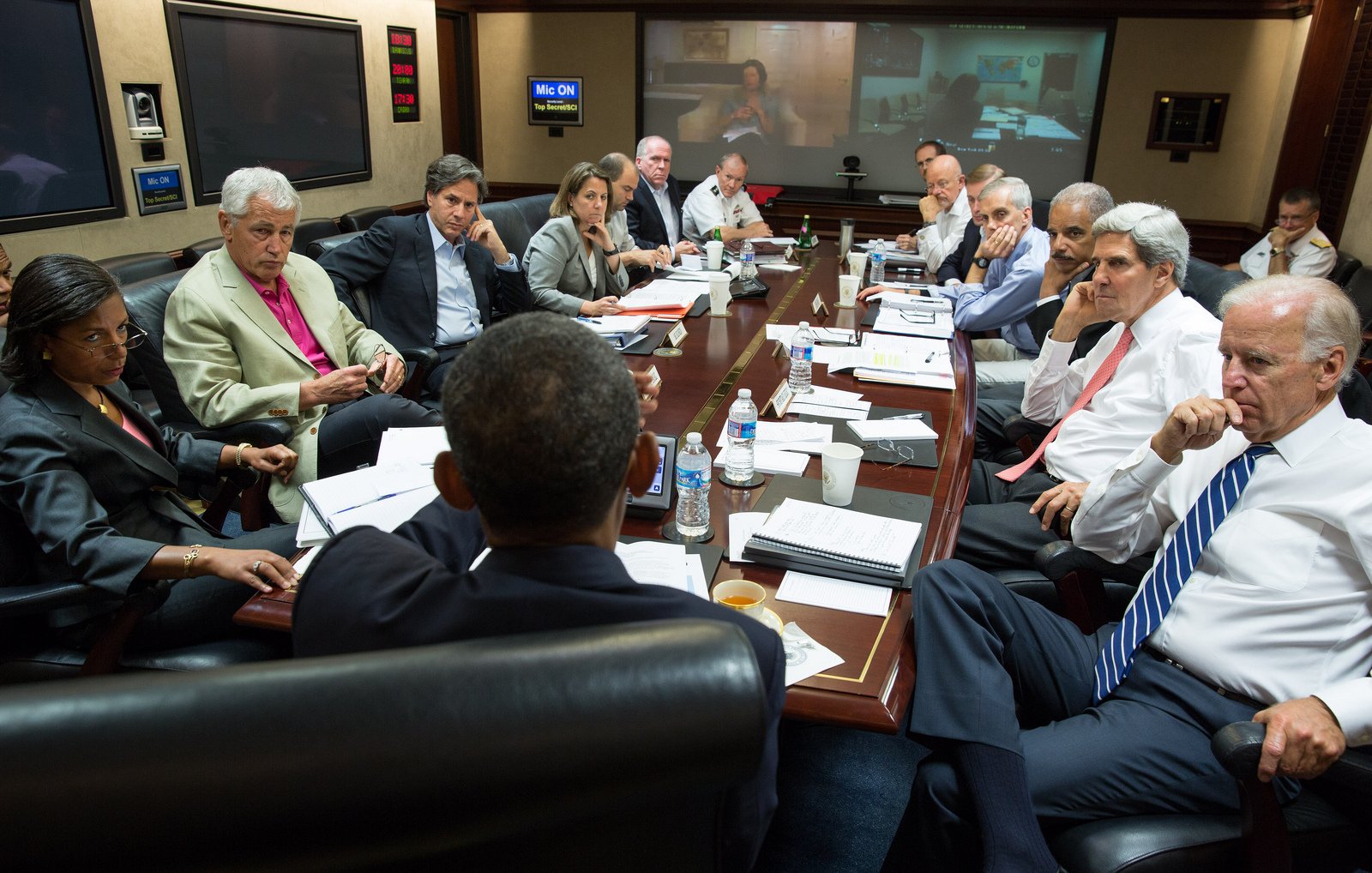 L'image montre une réunion importante autour d'une grande table. Plusieurs personnes, des hommes et des femmes, sont assis, engagés dans une discussion sérieuse. À l'arrière-plan, on peut voir un écran où se tient une vidéoconférence. Les participants semblent être des responsables et des conseillers, probablement dans un contexte gouvernemental ou militaire. L'ambiance générale est concentrée, avec des documents étalés sur la table et des bouteilles d'eau à disposition.