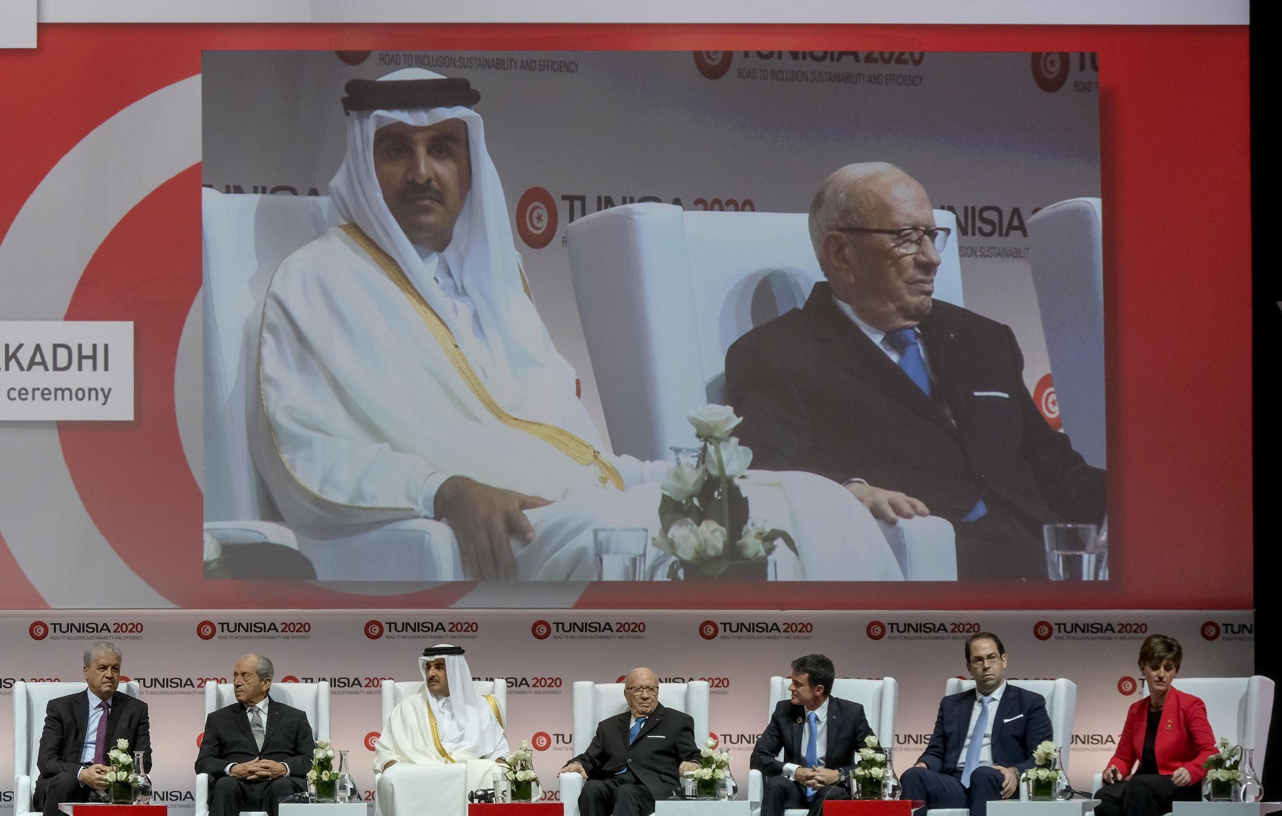 L'image montre une scène de conférence ou de séminaire avec plusieurs intervenants assis sur des chaises, sur une scène décorée avec des fleurs blanches. Au centre, il y a un grand écran affichant un homme en costume, vraisemblablement un orateur ou un participant à l'événement. Les participants semblent représentés sur une plateforme, prenant part à une discussion ou à une cérémonie. Le fond est décoré avec des motifs et des logos liés à l'événement "TUNISIA 2020". L'ambiance semble professionnelle et formelle.