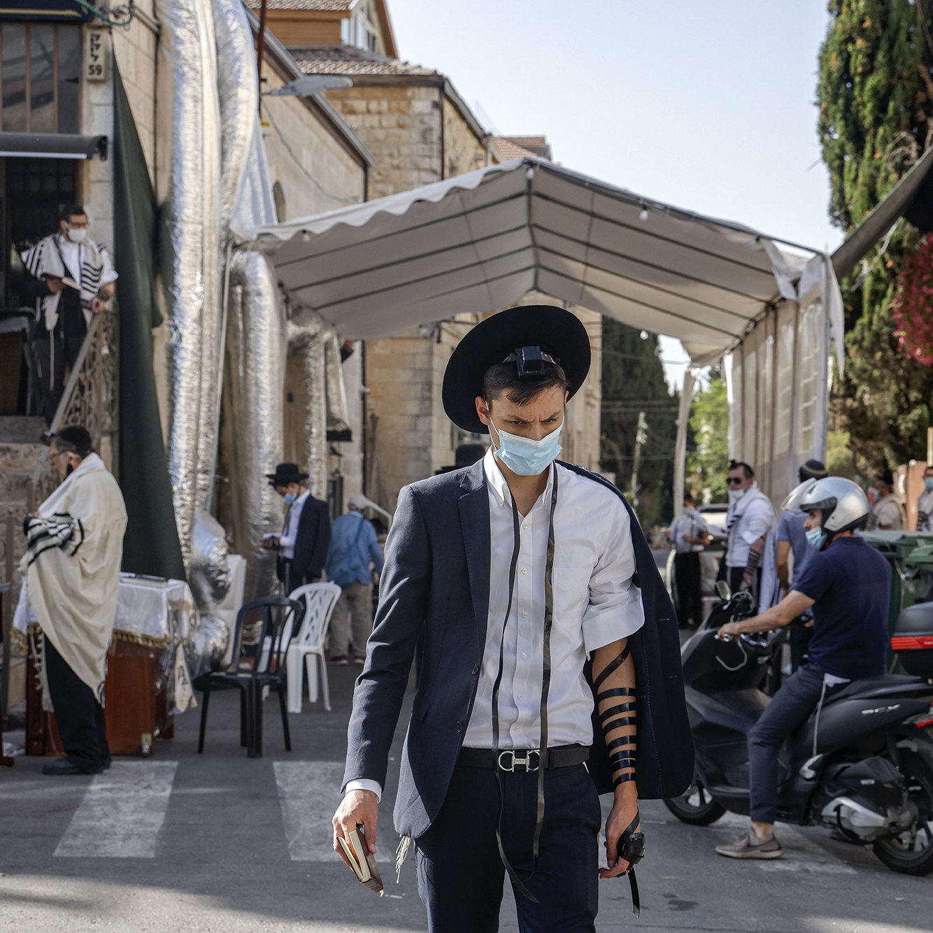 En la imagen se puede ver a un hombre joven que lleva puesta una camisa blanca y un saco oscuro, además de un sombrero negro. Está caminando por una calle que parece estar cerca de un lugar de culto judío, ya que al fondo se observa un edificio con elementos relacionados con la cultura y religión judía. El hombre lleva una máscara, posiblemente debido a las precauciones sanitarias. Se puede notar que tiene filacterias (tefilín) en su brazo, lo cual es parte de la práctica judía durante las oraciones. A su alrededor hay otras personas, algunas en la acera y otras en vehículos, lo que sugiere que es un lugar activo. Hay también un toldo montado, probablemente utilizado para eventos religiosos.