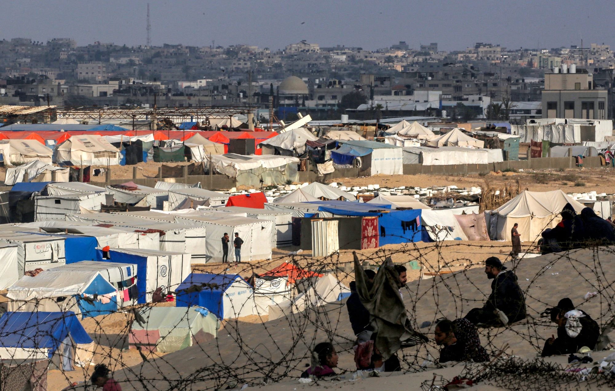L'image montre un camp de réfugiés avec de nombreuses tentes de différentes couleurs, principalement blanches et bleues, s'étendant sur un terrain désertique. À l'arrière-plan, on aperçoit une ville avec des bâtiments. On peut également distinguer des personnes assises, ainsi que des barbelés entourant le camp, ce qui souligne une ambiance de sécurité précaire et d'incertitude. L'ensemble évoque une situation difficile et des conditions de vie sommaires.