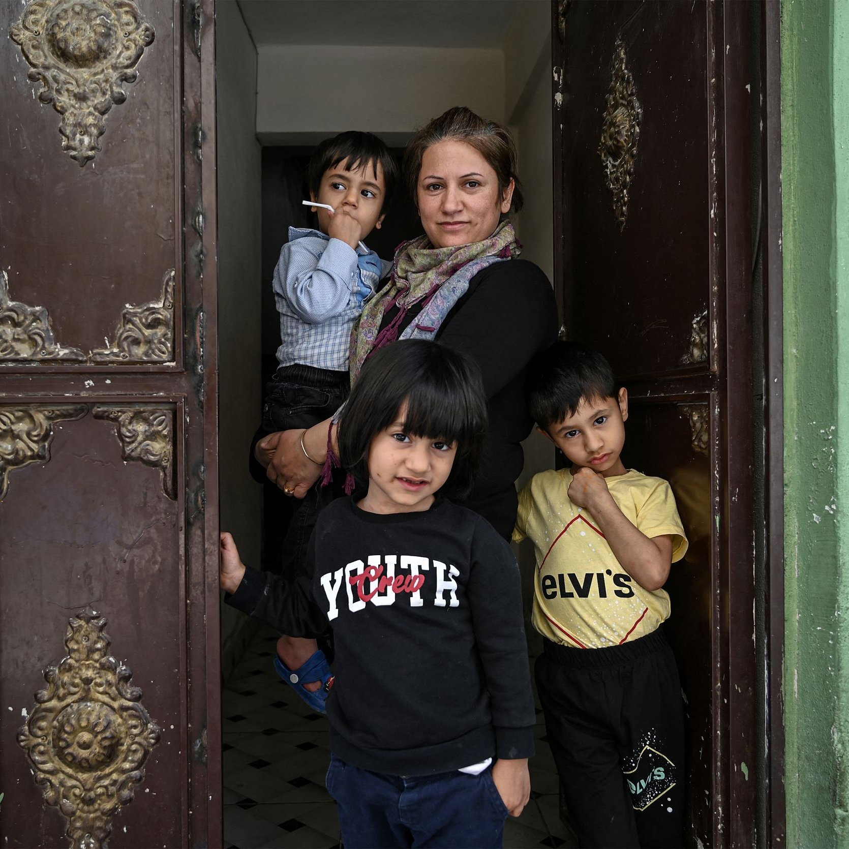 L'image montre une femme debout dans une porte d'entrée, entourée de trois jeunes enfants. Elle a un air calme et tient une cigarette. Les enfants semblent curieux et sont vêtus de vêtements décontractés. Leurs expressions varient, certains souriant tandis que d'autres regardent vers l'objectif. Le fond présente des murs verts et une porte ornée de détails décoratifs, ajoutant une touche particulière à la scène.