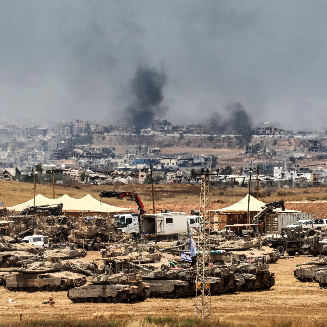 Terreno desolato con carri armati, fumi di incendi e un paesaggio urbano distrutto sullo sfondo.