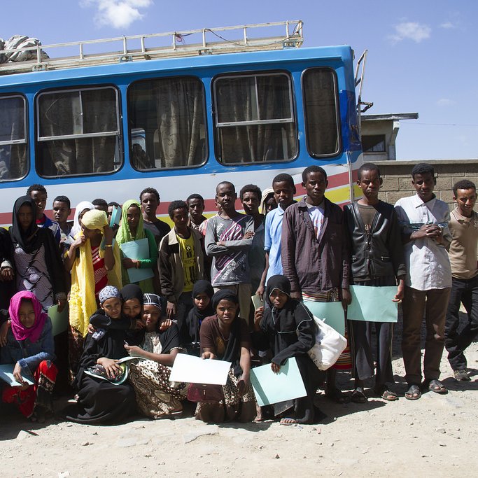 L'image montre un groupe de jeunes adultes et d'adolescents rassemblés devant un bus coloré. Ils semblent être en train de poser pour la photo, tenant tous des documents ou des fichiers. Les personnes portent des vêtements variés, incluant des hijabs et des tenues traditionnelles. L'environnement semble être en plein air, avec un ciel clair et quelques structures visibles en arrière-plan. L'atmosphère paraît conviviale et joyeuse.