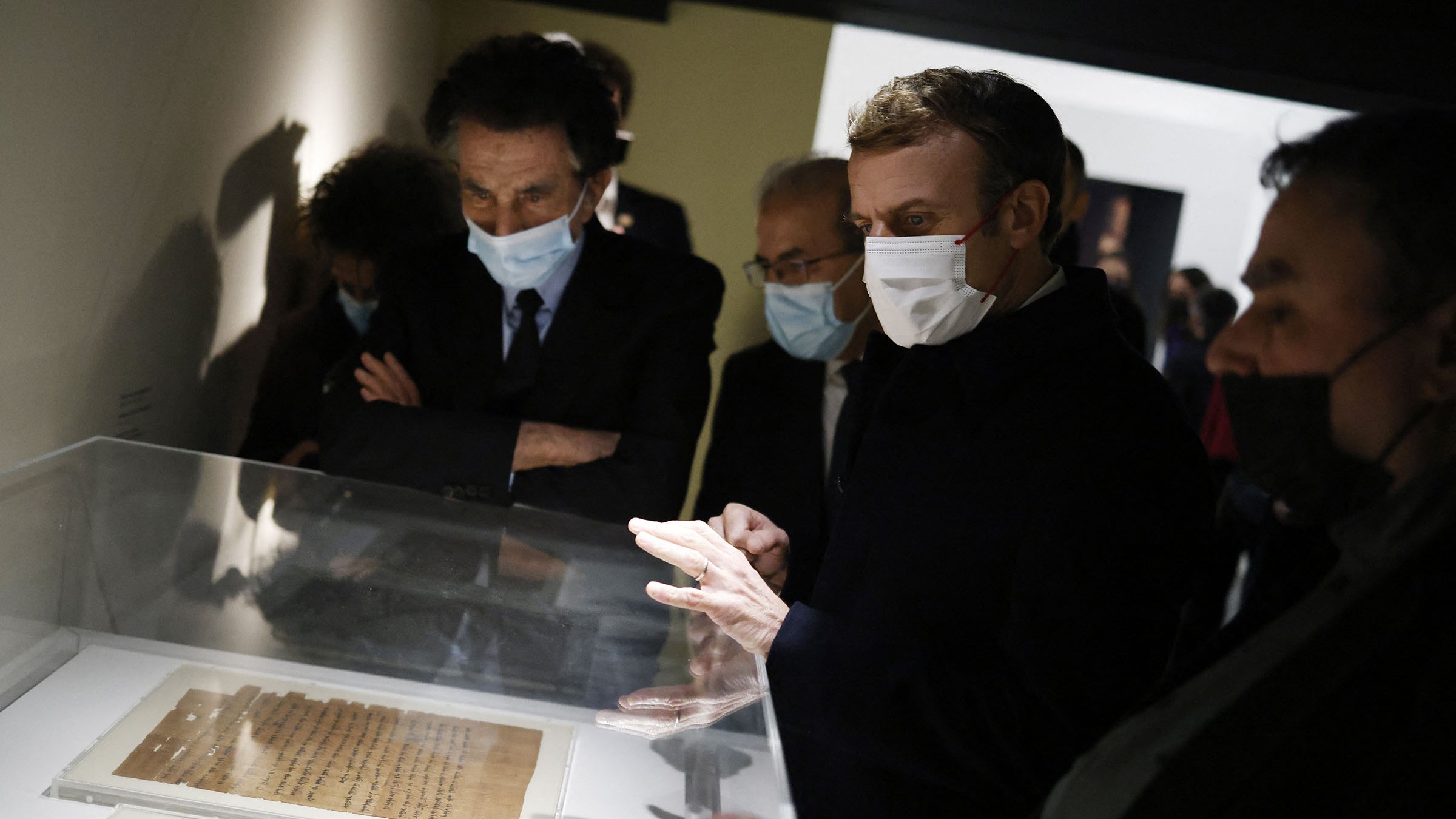 تظهر الصورة مجموعة من الأشخاص يتفحصون وثائق تاريخية محفوظة في علبة زجاجية. يبدو أن بينهم رجل يرتدي بدلة سوداء وقناع وجه، يُعبر عن اهتمامه بالمعروضات. الأشخاص الآخرون يرتدون أيضاً كمامات ويتحدثون مع بعضهم البعض. الخلفية محاطة بإضاءة خافتة، مما يعطي انطباعًا جادًا ومناسبًا لعرض آثار ثقافية.