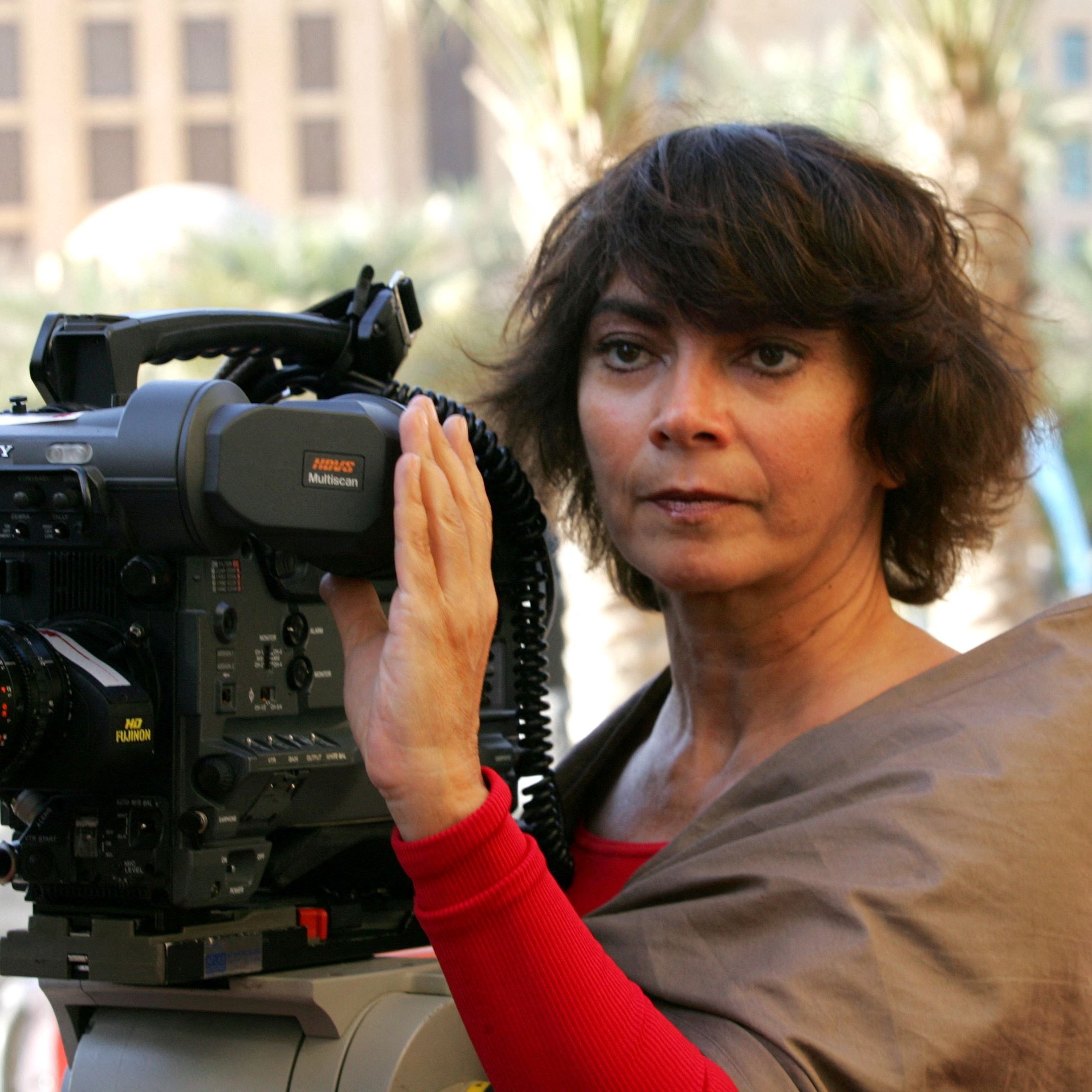 L'image montre une femme debout devant une caméra vidéo professionnelle. Elle a les cheveux bruns et bouclés et porte un haut de couleur claire. La femme semble concentrée, en train de préparer ou d'utiliser la caméra, qui est positionnée sur un trépied. En arrière-plan, on peut voir des éléments d'architecture moderne. L'éclairage semble naturel, suggérant que la scène se déroule à l'extérieur.